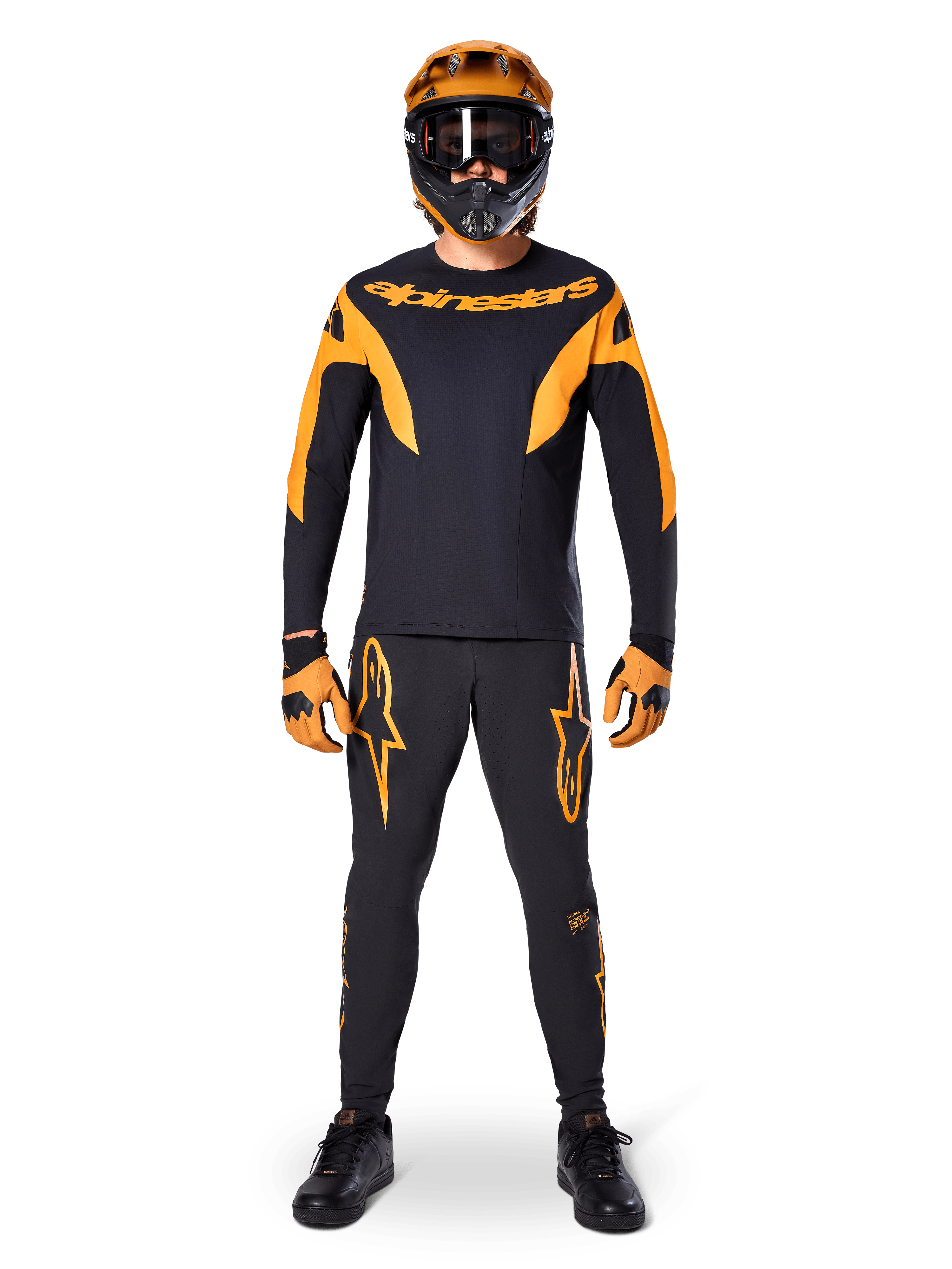 Pantalones Alpinestars A-Supra Race, pantalones de ciclismo de montaña, negros y naranjas, usados por un piloto con un equipamiento de carreras completo que incluye jersey negro y naranja, casco naranja y guantes a juego, con llamativos logotipos de estrella naranjas en las piernas