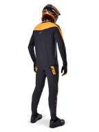 Pantalones Alpinestars A-Supra Race, pantalones de competición MTB, negros con logotipos naranjas, vistos desde atrás en un ciclista con jersey, casco y guantes negros y naranjas a juego, resaltando el logotipo inspirado en MX en el canesú trasero y el ajuste de rendimiento para senderos técnicos.