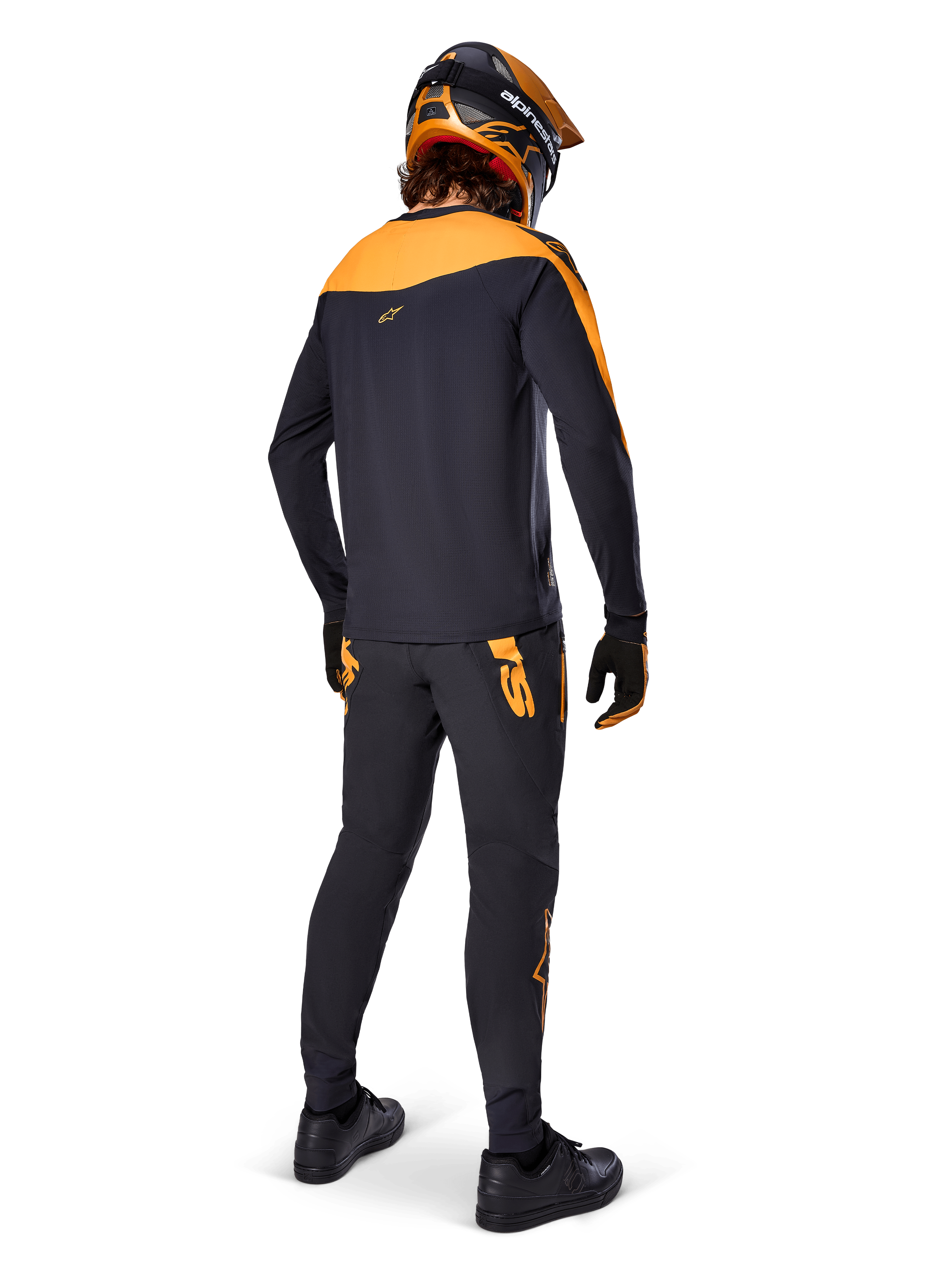 Pantalones Alpinestars A-Supra Race, pantalones de competición MTB, negros con logotipos naranjas, vistos desde atrás en un ciclista con jersey, casco y guantes negros y naranjas a juego, resaltando el logotipo inspirado en MX en el canesú trasero y el ajuste de rendimiento para senderos técnicos.