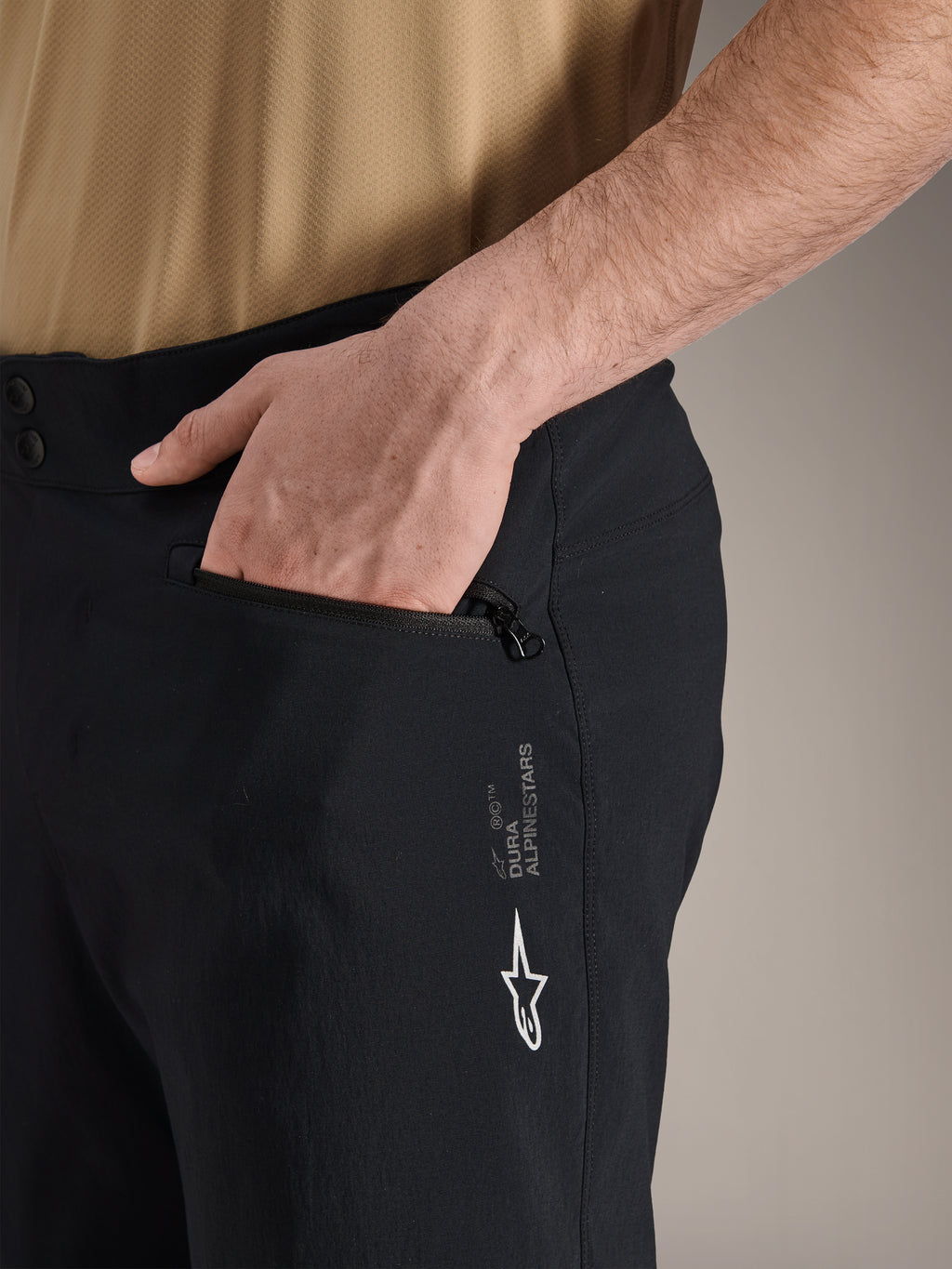 Alpinestars A-Dura Shorts, shorts de ciclismo de montaña negros, primer plano de bolsillo lateral con cremallera con una mano en el interior, con el logotipo blanco de Alpinestars y la marca A-Dura, tejido textil duradero para ciclismo todoterreno