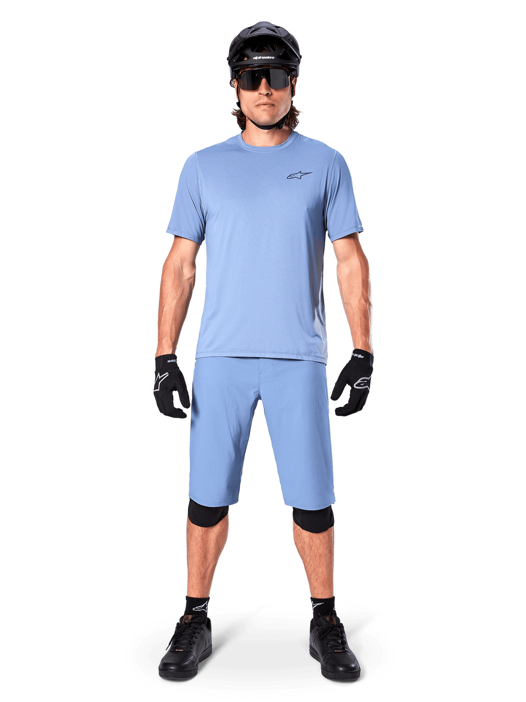 Modelo con los shorts Alpinestars A-Dura en color Infinity Blue, jersey azul claro a juego, casco de bicicleta de montaña negro, gafas de sol negras, guantes y rodilleras negros, equipamiento completo de MTB para trail