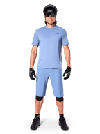 Modelo con los shorts Alpinestars A-Dura en color Infinity Blue, jersey azul claro a juego, casco de bicicleta de montaña negro, gafas de sol negras, guantes y rodilleras negros, equipamiento completo de MTB para trail