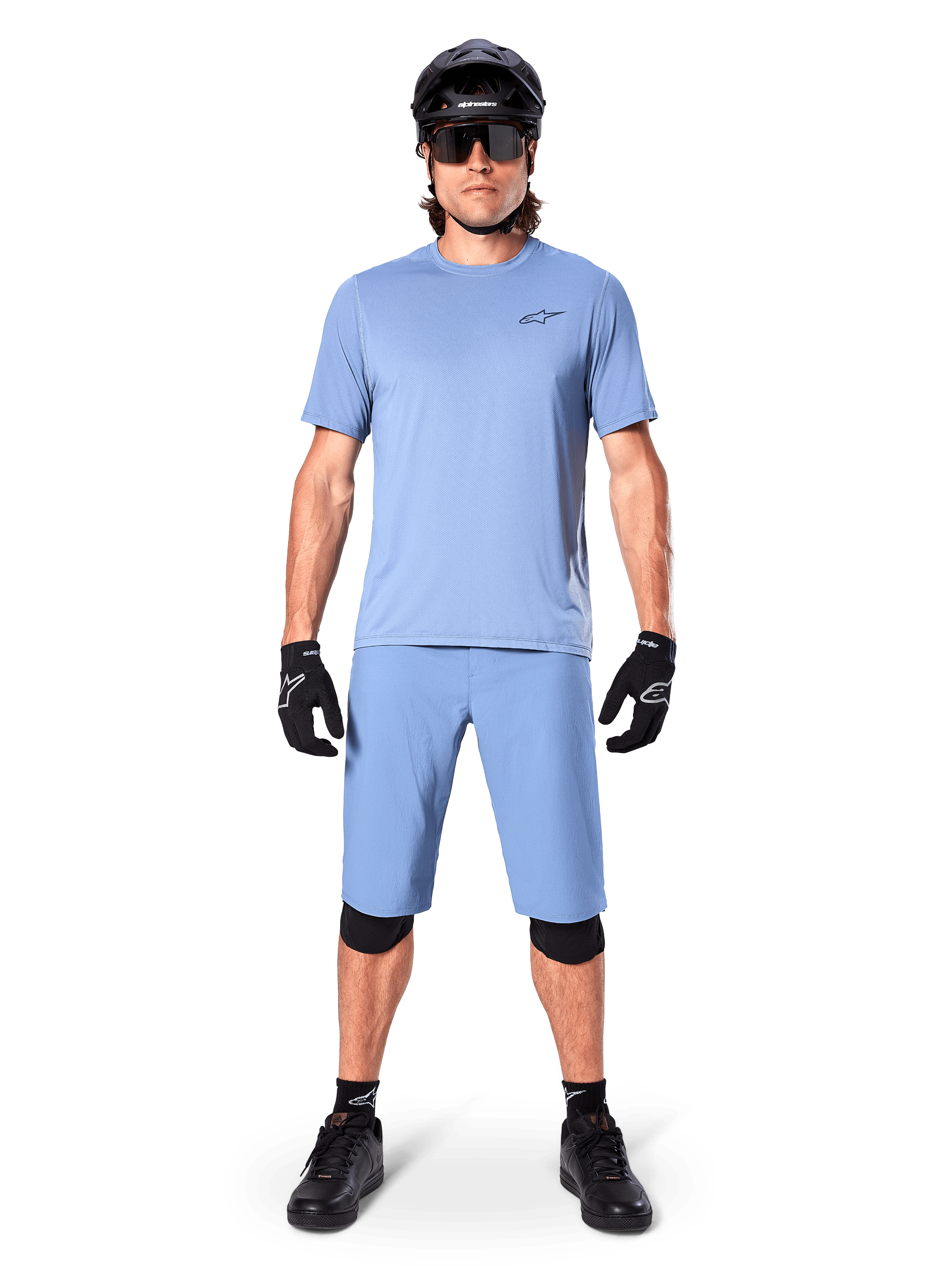Modelo con los shorts Alpinestars A-Dura en color Infinity Blue, jersey azul claro a juego, casco de bicicleta de montaña negro, gafas de sol negras, guantes y rodilleras negros, equipamiento completo de MTB para trail