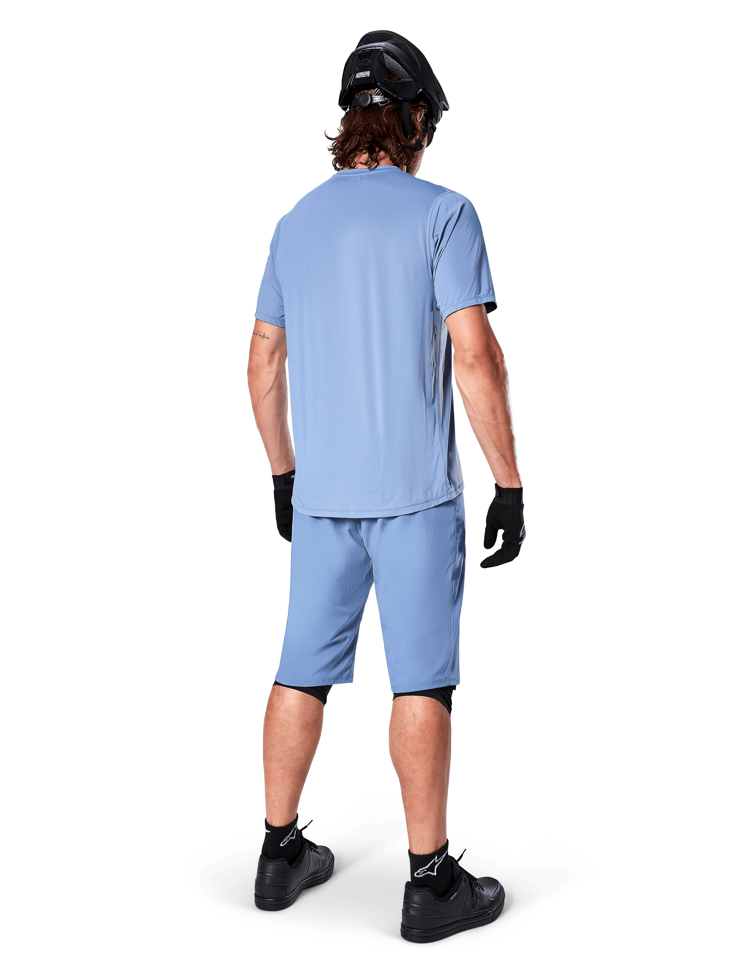 Vista trasera de un ciclista de montaña con shorts Alpinestars A-Dura en color Infinity Blue, jersey de manga corta a juego, casco negro, guantes negros y zapatillas negras, equipamiento coordinado de alto rendimiento para trail MTB