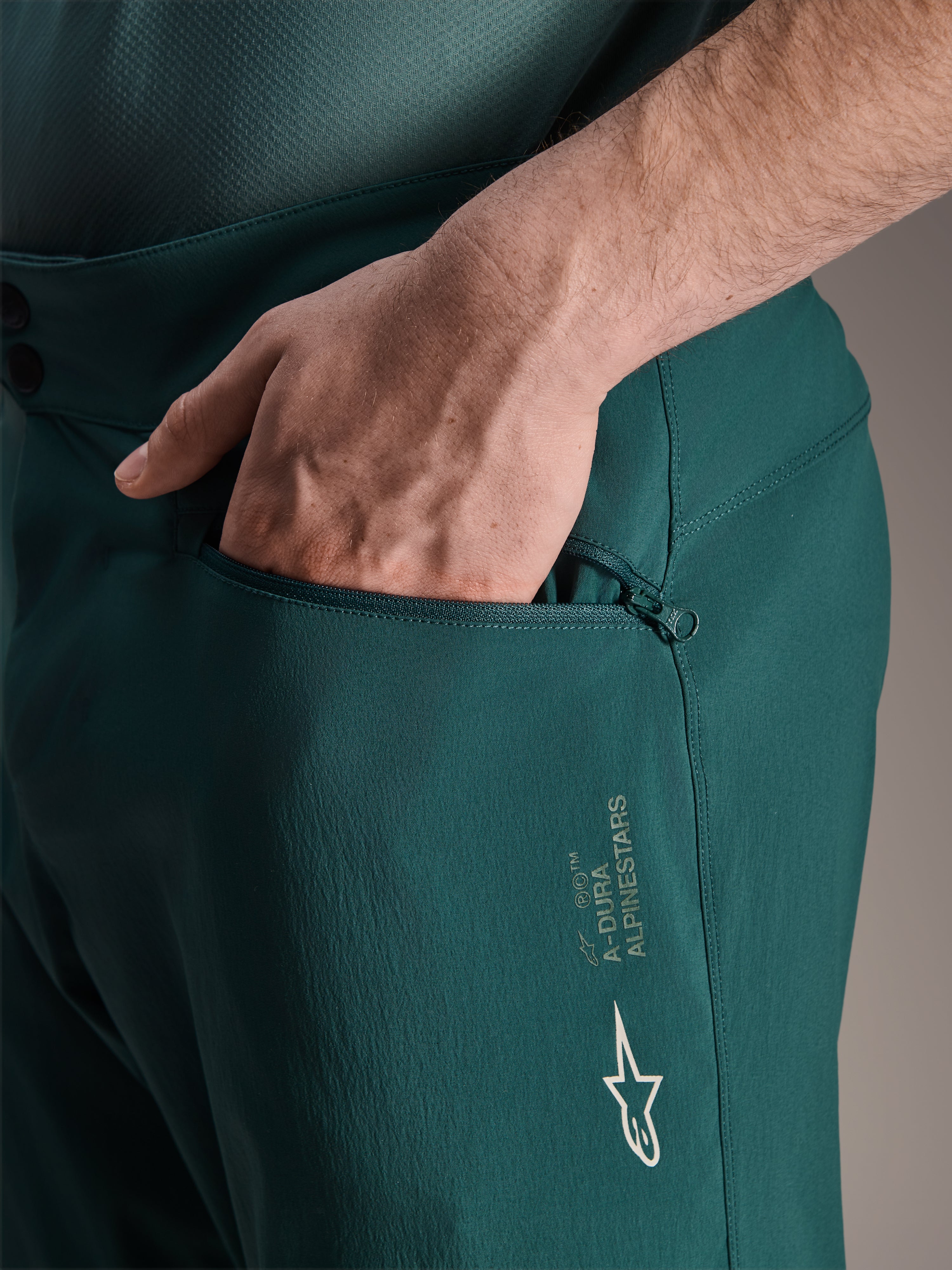 Shorts Alpinestars A-Dura en Dark Pine, primer plano que muestra una mano en el bolsillo con cremallera y forro de malla, con el logo de la estrella blanca de Alpinestars y la marca A-Dura, tejido resistente para ciclismo de montaña