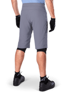 Shorts Alpinestars A-Dura, shorts de mountain bike en color gris oscuro, vista trasera mostrada en un ciclista, usados con guantes negros, mallas para rodillas negras y calcetines negros con el logo de Alpinestars, construcción duradera para ciclismo de montaña