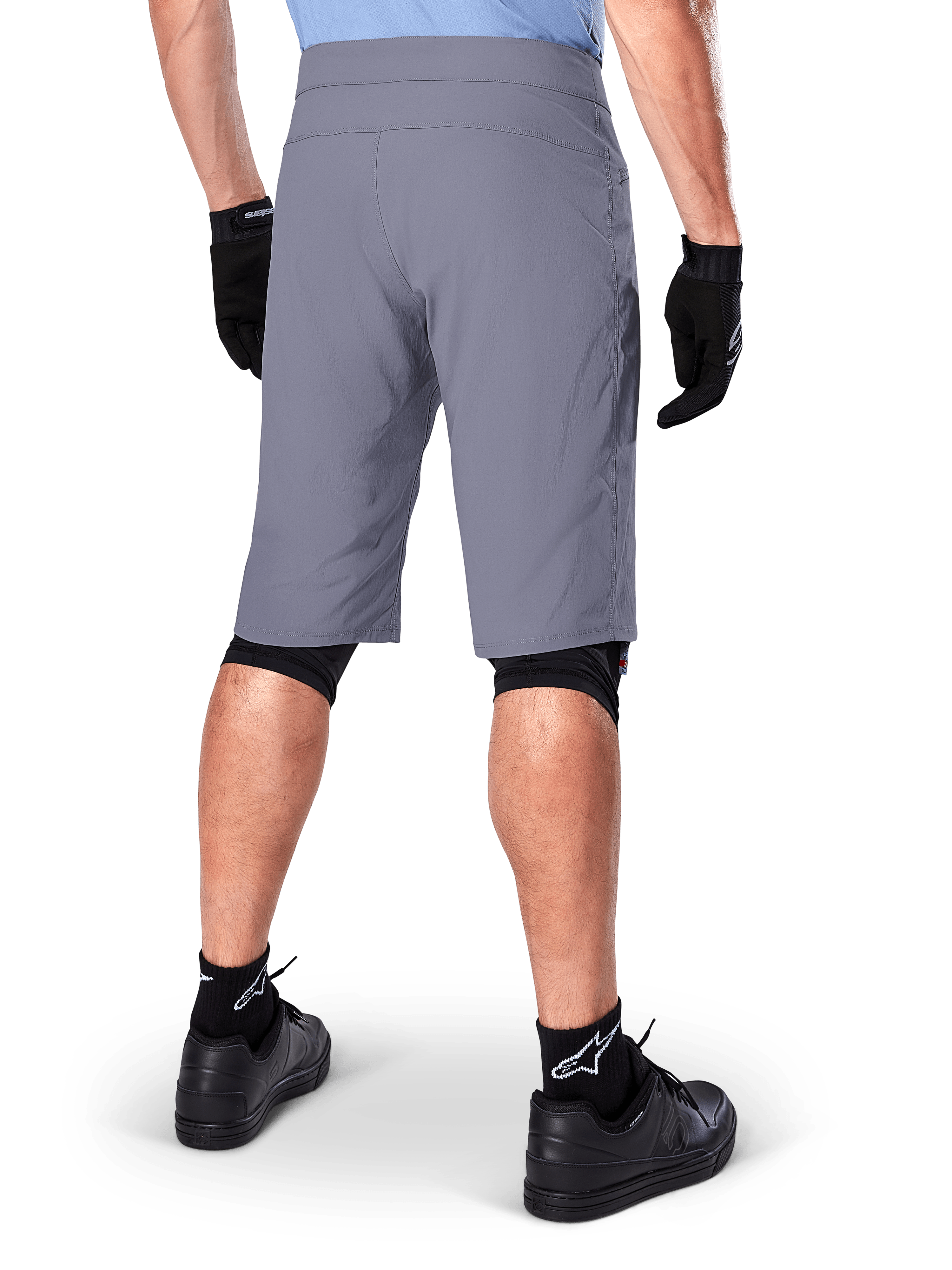 Shorts Alpinestars A-Dura, shorts de mountain bike en color gris oscuro, vista trasera mostrada en un ciclista, usados con guantes negros, mallas para rodillas negras y calcetines negros con el logo de Alpinestars, construcción duradera para ciclismo de montaña