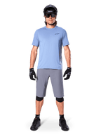 Ciclista masculino con Alpinestars A-Dura Shorts en gris oscuro, combinados con una camiseta de manga corta azul claro, casco negro, gafas de sol y rodilleras negras, equipamiento completo de ciclismo de montaña para rendimiento off-road