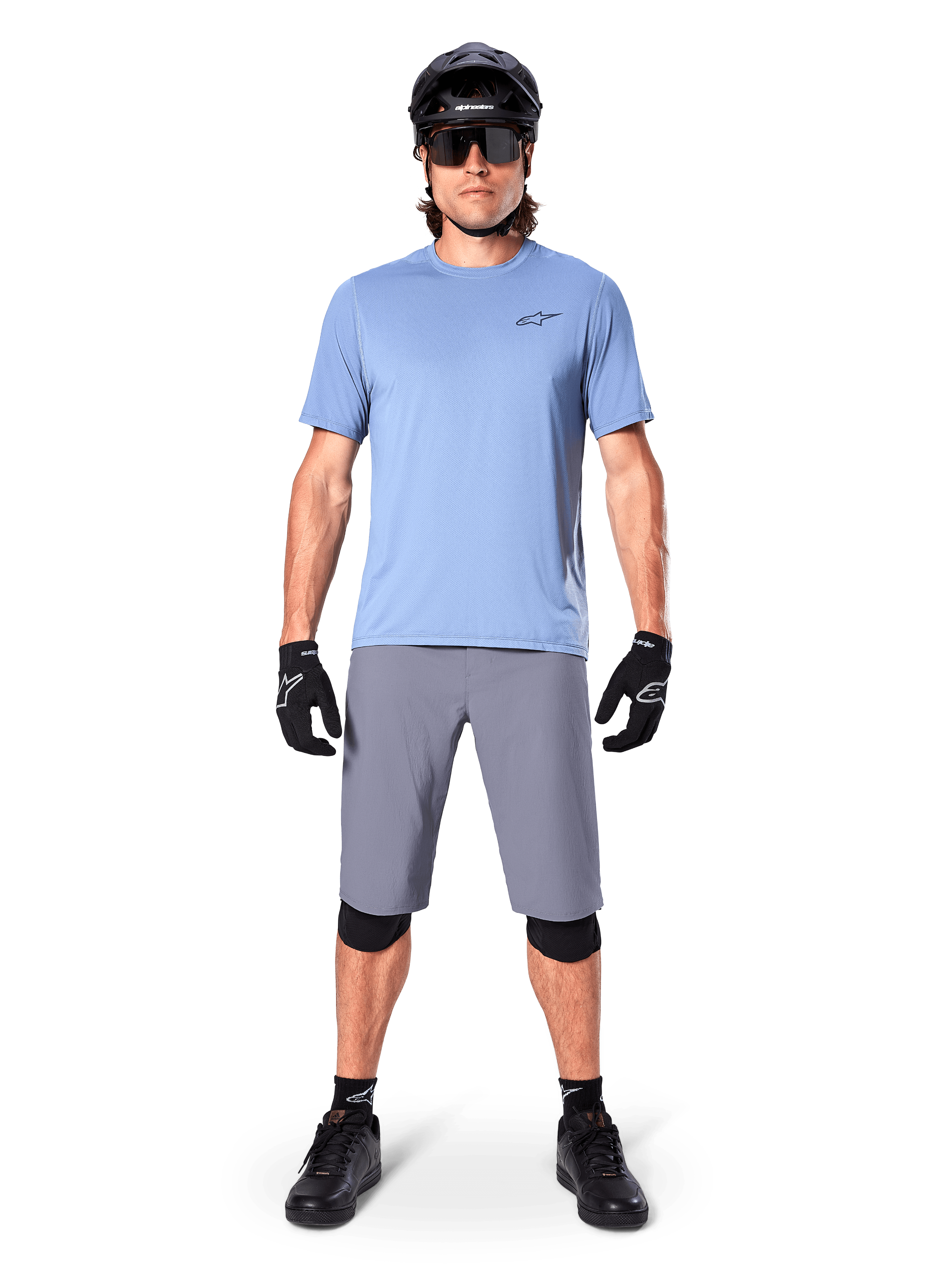 Ciclista masculino con Alpinestars A-Dura Shorts en gris oscuro, combinados con una camiseta de manga corta azul claro, casco negro, gafas de sol y rodilleras negras, equipamiento completo de ciclismo de montaña para rendimiento off-road