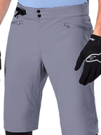 Alpinestars A-Dura Shorts, shorts de MTB, gris oscuro, vista de cerca de la cintura que muestra el cierre de doble broche, ajustador lateral de cintura y bolsillos con cremallera, combinados con guantes de ciclismo negros, ropa duradera para ciclismo de montaña.
