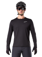 Jersey Térmico Alpinestars A-Dura, jersey de ciclismo de montaña de manga larga en color negro, usado con casco negro y gafas de sol grandes, con un pequeño logotipo blanco de Alpinestars en el pecho, ajuste regular, ropa de ciclismo MTB