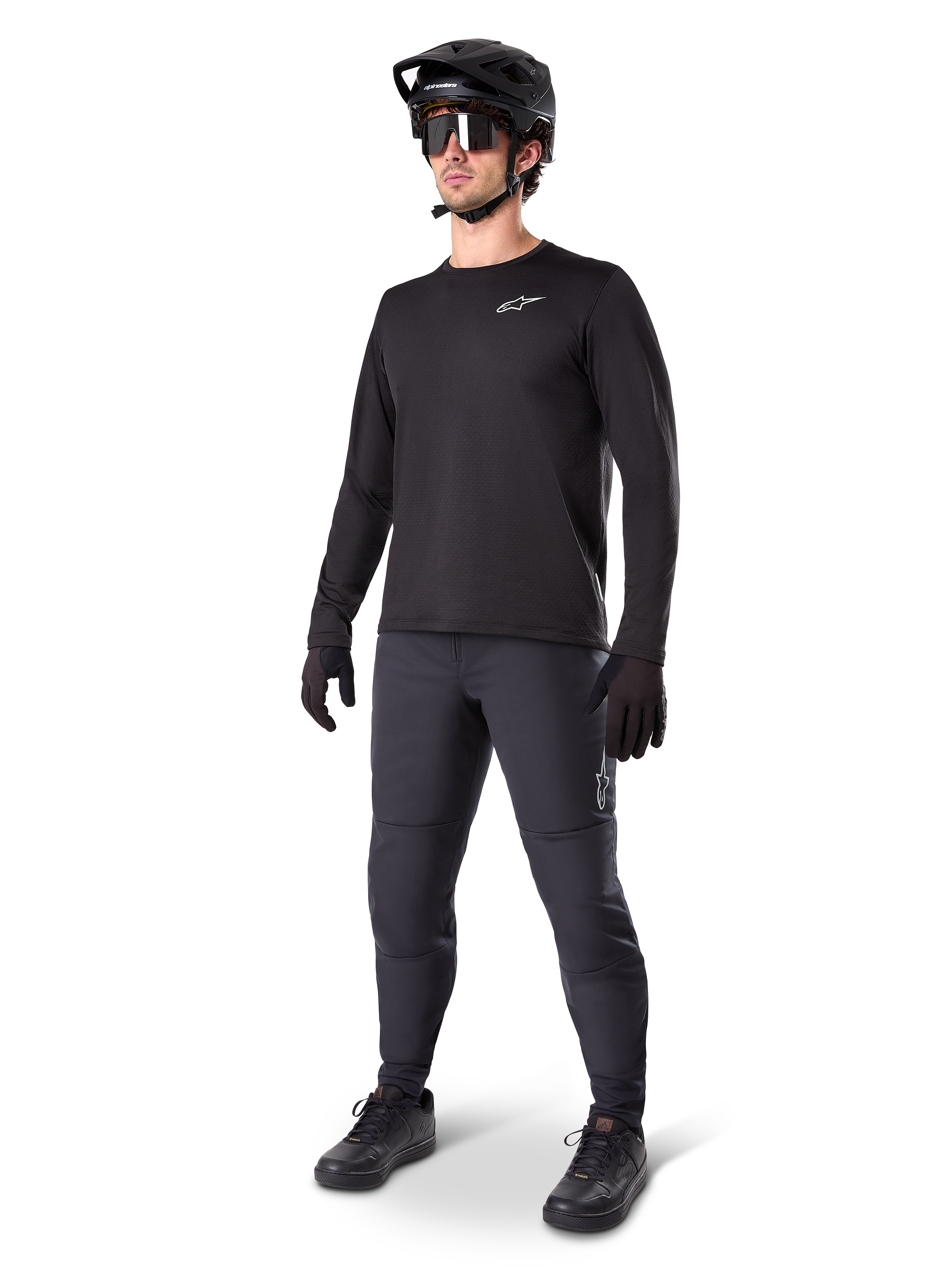 Modelo con el Jersey Térmico Alpinestars A-Dura, jersey de MTB de manga larga negro, casco de MTB negro, gafas de sol y pantalones gris oscuro, con un pequeño logotipo blanco en el pecho y forro polar aislante para montar en climas fríos.