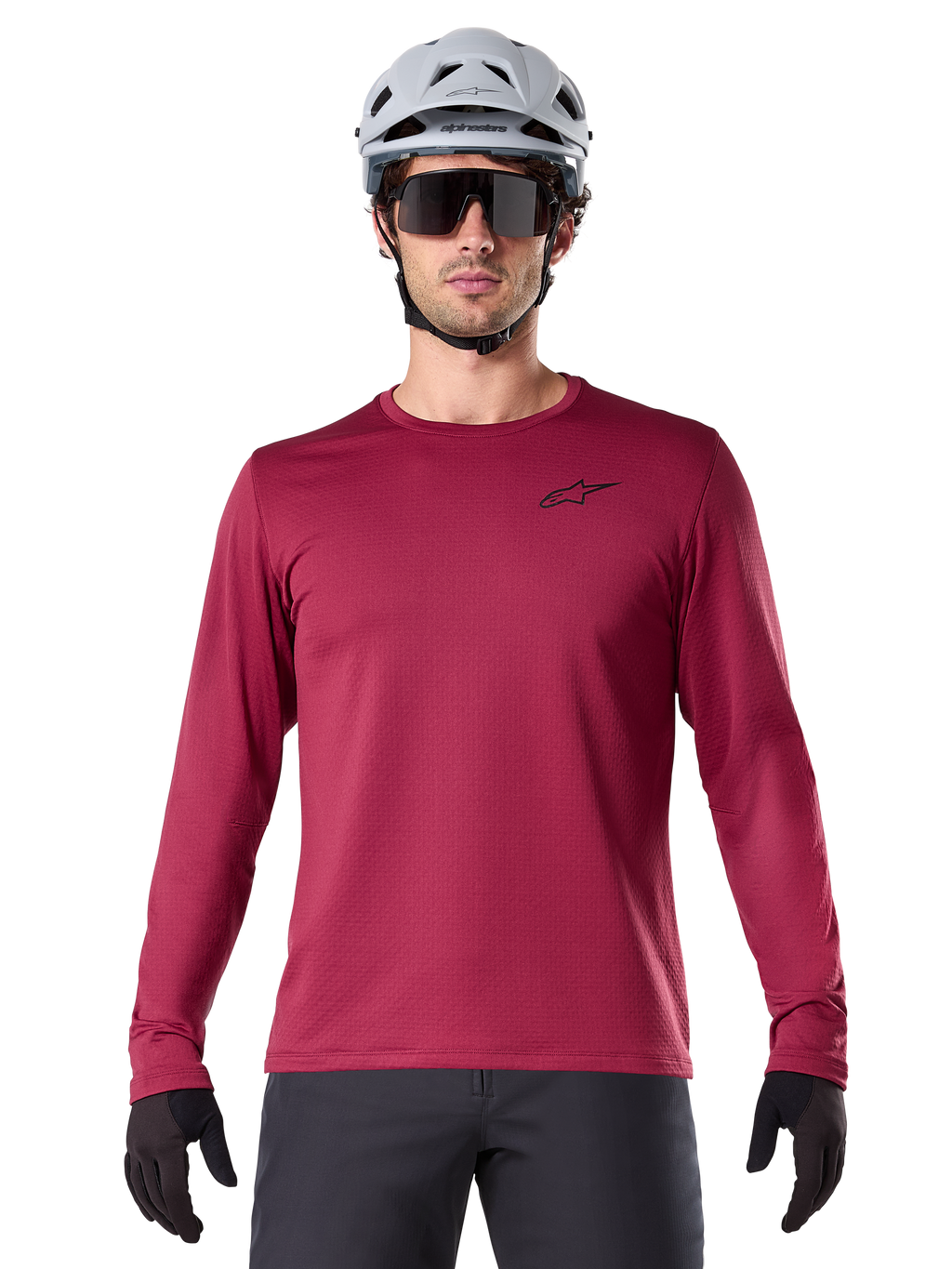 Ciclista masculino con el Jersey Térmico A-Dura de Alpinestars, jersey de MTB de manga larga, color granate, combinado con un casco gris y gafas de sol negras, presenta un pequeño logotipo de estrella negro en el pecho y tejido de vellón aislante para ciclismo de montaña en climas fríos