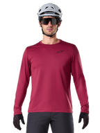 Ciclista masculino con el Jersey Térmico A-Dura de Alpinestars, jersey de MTB de manga larga, color granate, combinado con un casco gris y gafas de sol negras, presenta un pequeño logotipo de estrella negro en el pecho y tejido de vellón aislante para ciclismo de montaña en climas fríos