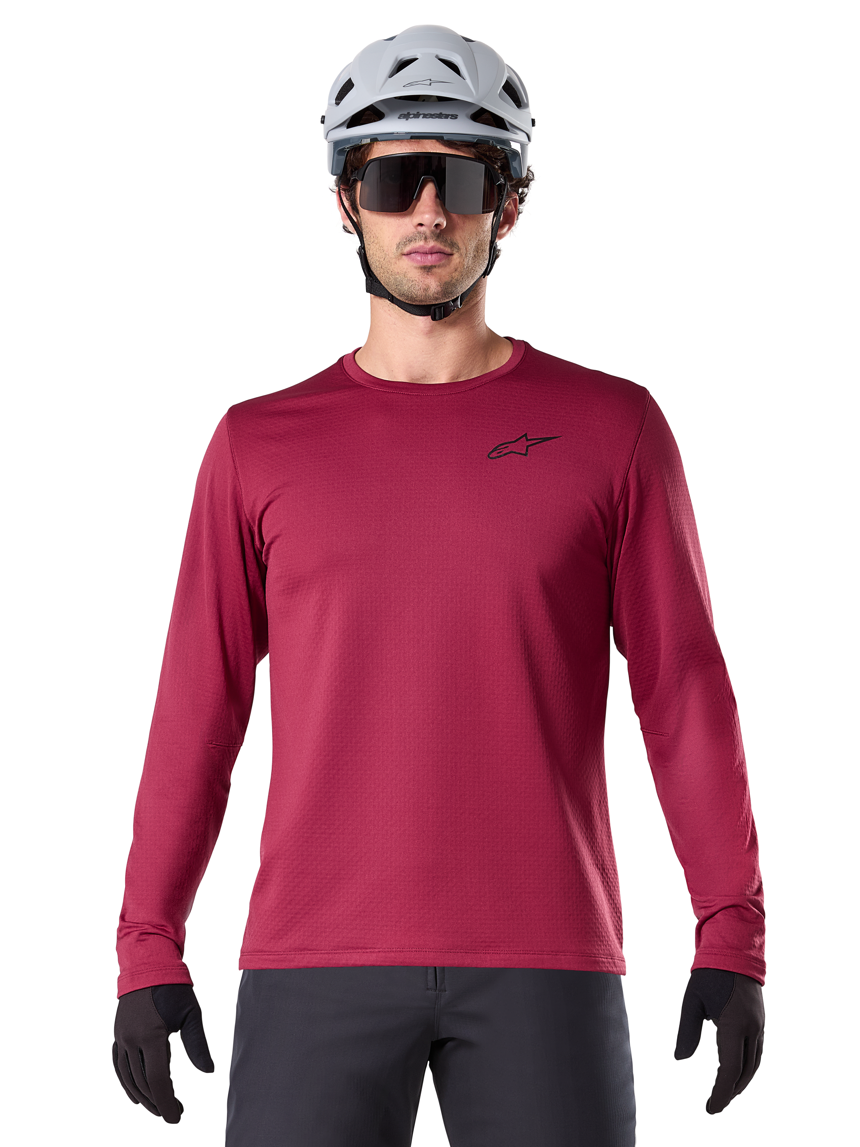 Ciclista masculino con el Jersey Térmico A-Dura de Alpinestars, jersey de MTB de manga larga, color granate, combinado con un casco gris y gafas de sol negras, presenta un pequeño logotipo de estrella negro en el pecho y tejido de vellón aislante para ciclismo de montaña en climas fríos