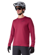 Alpinestars A-Dura Thermal Jersey, jersey de manga larga para ciclismo de montaña, rojo borgoña, con forro polar aislante y un pequeño logotipo negro en el pecho, usado con un casco gris, gafas de sol negras y guantes negros, diseñado para ciclismo en climas fríos
