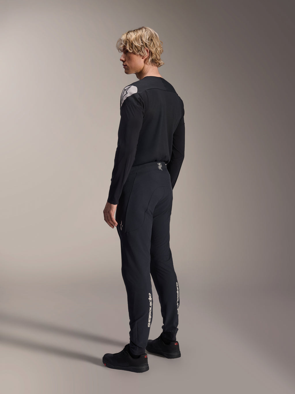 Pantalones Alpinestars A-Aria Elite, pantalones de MTB, Black Titanium, vista trasera en tres cuartos en modelo, con logotipo de Alpinestars en blanco en la parte inferior de las piernas, ajuste de rendimiento con puños elásticos, combinados con un jersey de manga larga negro a juego.