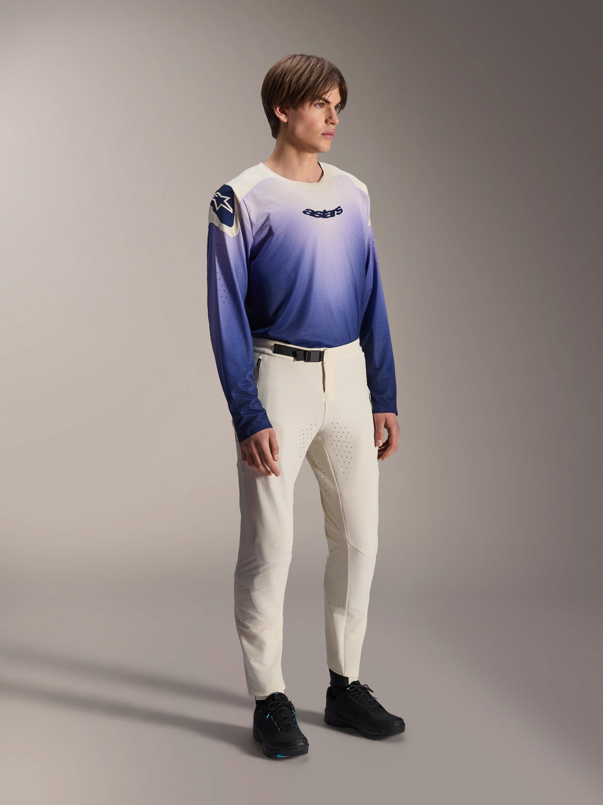 Modelo masculino con el jersey Alpinestars A-Aria Polartec Maker Elite en color Baja Blue, con diseño degradado de blanco a azul y logotipo 'astars' negro en el pecho, combinado con pantalones de MTB blanco roto y zapatillas negras, ropa técnica de alto rendimiento para trail.