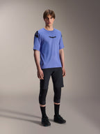 Modelo masculino con la camiseta de manga corta Alpinestars A-Aria Polartec Elite en color Baja Blue, combinada con pantalones cortos de MTB y rodilleras negros, con logotipos Astars negros en el pecho y el hombro, equipamiento de alto rendimiento para bicicleta de montaña