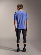 Modelo masculino visto desde atrás con equipamiento Alpinestars para ciclismo de montaña, jersey de manga corta A-Aria Polartec Elite azul claro, shorts negros A-Aria Elite, protectores de rodilla negros, calcetines negros y zapatillas negras, conjunto coordinado de alto rendimiento para MTB.
