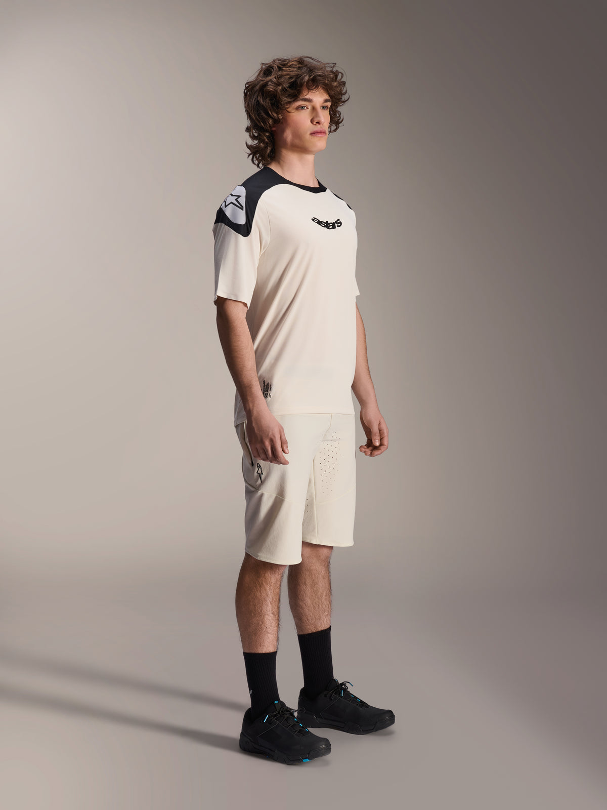 Modelo masculino con Shorts Alpinestars A-Aria Elite en color Ecru, shorts técnicos de mountain bike con ventilación cortada con láser, combinados con un jersey de manga corta a juego en crema y negro, y zapatillas negras, ropa de ciclismo profesional