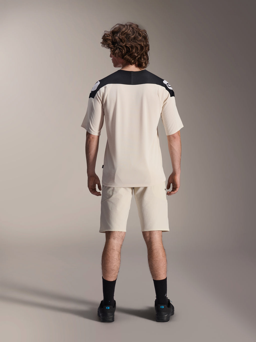 Alpinestars A-Aria Elite Shorts y Jersey A-Aria Polartec Elite de manga corta, color crudo con paneles negros en los hombros, vista trasera de un modelo con equipamiento de alto rendimiento para bicicleta de montaña, combinado con calcetines y zapatillas negros