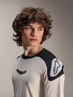 Camiseta de manga corta Alpinestars A-Aria Polartec Elite, color crudo con paneles negros en los hombros, con el logotipo astars negro en el pecho y el logotipo Astars blanco en el hombro, equipamiento de alto rendimiento para bicicleta de montaña usado por un modelo