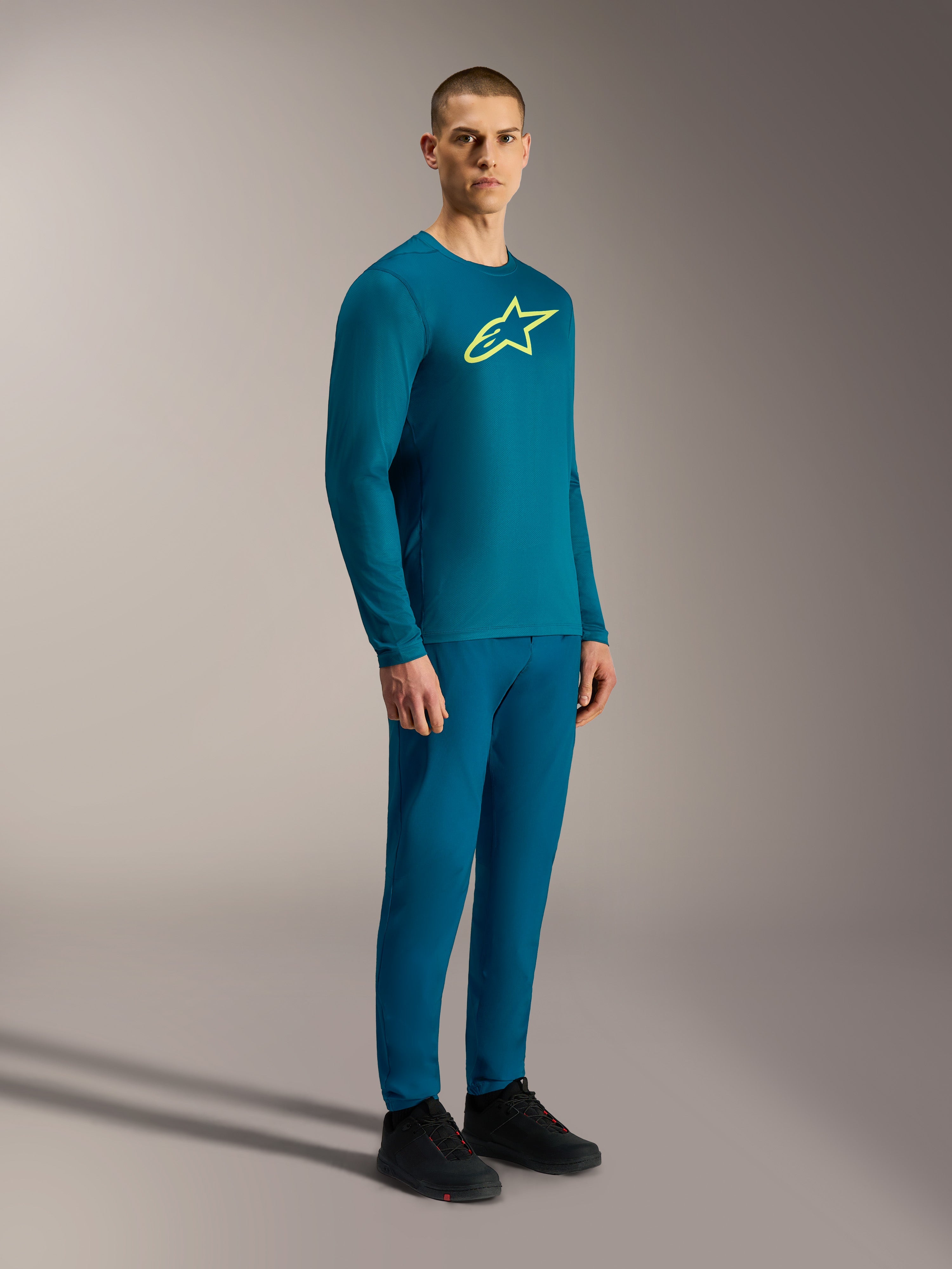 Camiseta A-Dura Ride de manga larga azul verde con logo Astars amarillo, conjunto de pantalones MTB y calzado técnico.