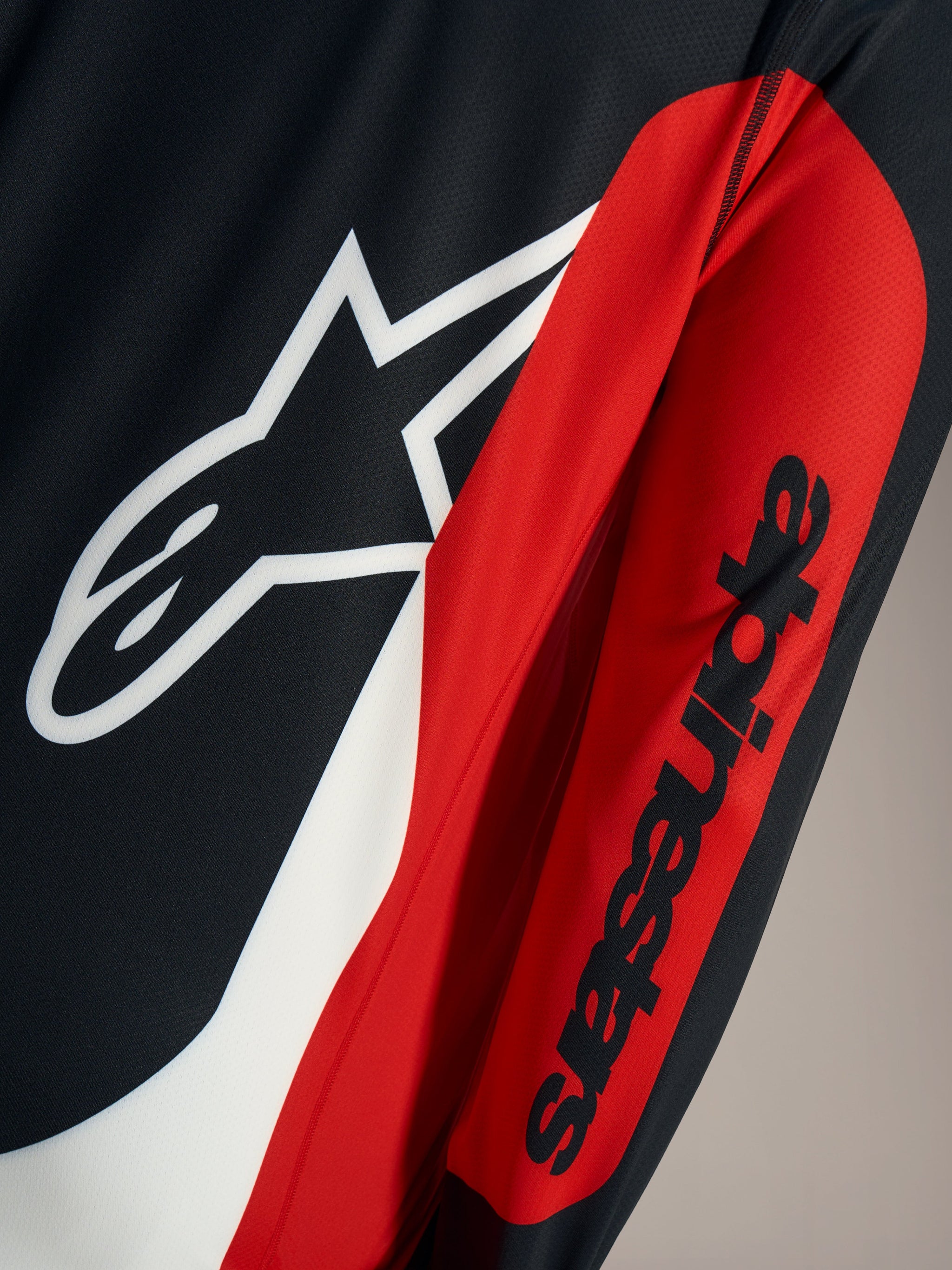 Camiseta A-Dura Race, detalle del logotipo Astars blanco y marca Alpinestars negra sobre malla técnica roja y negra.