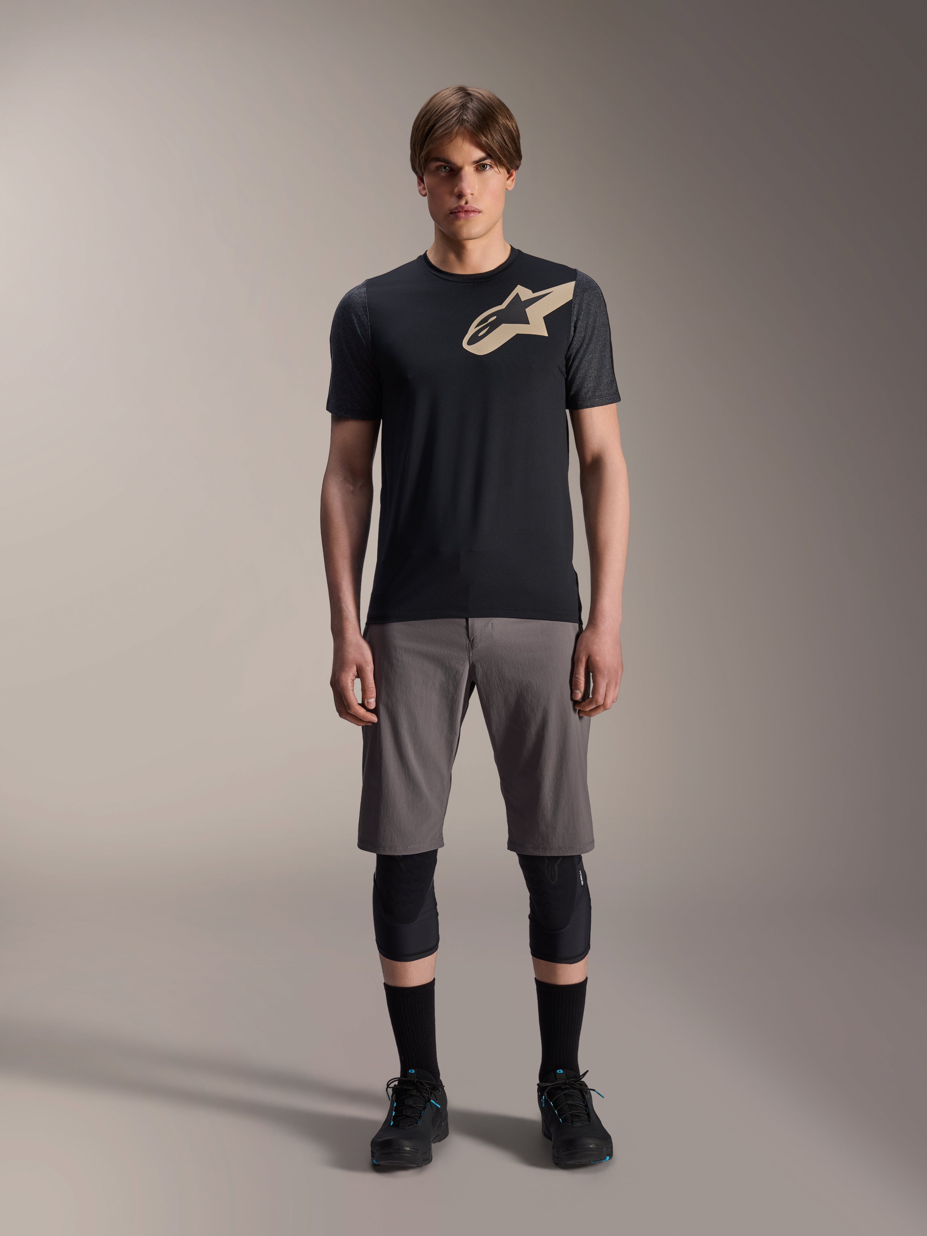 Jersey de manga corta Alpinestars A-Dura Lipan, negro con mangas gris jaspeado y logotipo Astars color canela en el pecho, usado por un modelo masculino con pantalones cortos de ciclismo de montaña grises y rodilleras negras, equipamiento técnico para MTB.