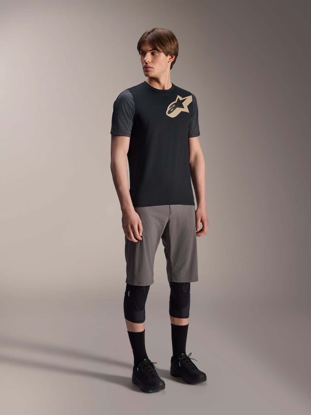 Modelo masculino luciendo el Jersey de manga corta Alpinestars A-Dura Lipan en color negro, con mangas gris oscuro y logotipo de Astars en color canela en el pecho, combinado con pantalones cortos grises de ciclismo de montaña, rodilleras negras y zapatillas de ciclismo negras, equipamiento coordinado de MTB