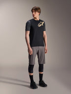 Modelo masculino luciendo el Jersey de manga corta Alpinestars A-Dura Lipan en color negro, con mangas gris oscuro y logotipo de Astars en color canela en el pecho, combinado con pantalones cortos grises de ciclismo de montaña, rodilleras negras y zapatillas de ciclismo negras, equipamiento coordinado de MTB