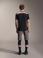Alpinestars A-Dura Lipan Jersey - Manga corta, negro con panel superior de la espalda en color tostado, vista trasera que muestra la cola más larga, usado con pantalones cortos de MTB grises y rodilleras negras, ropa de alto rendimiento para bicicleta de montaña