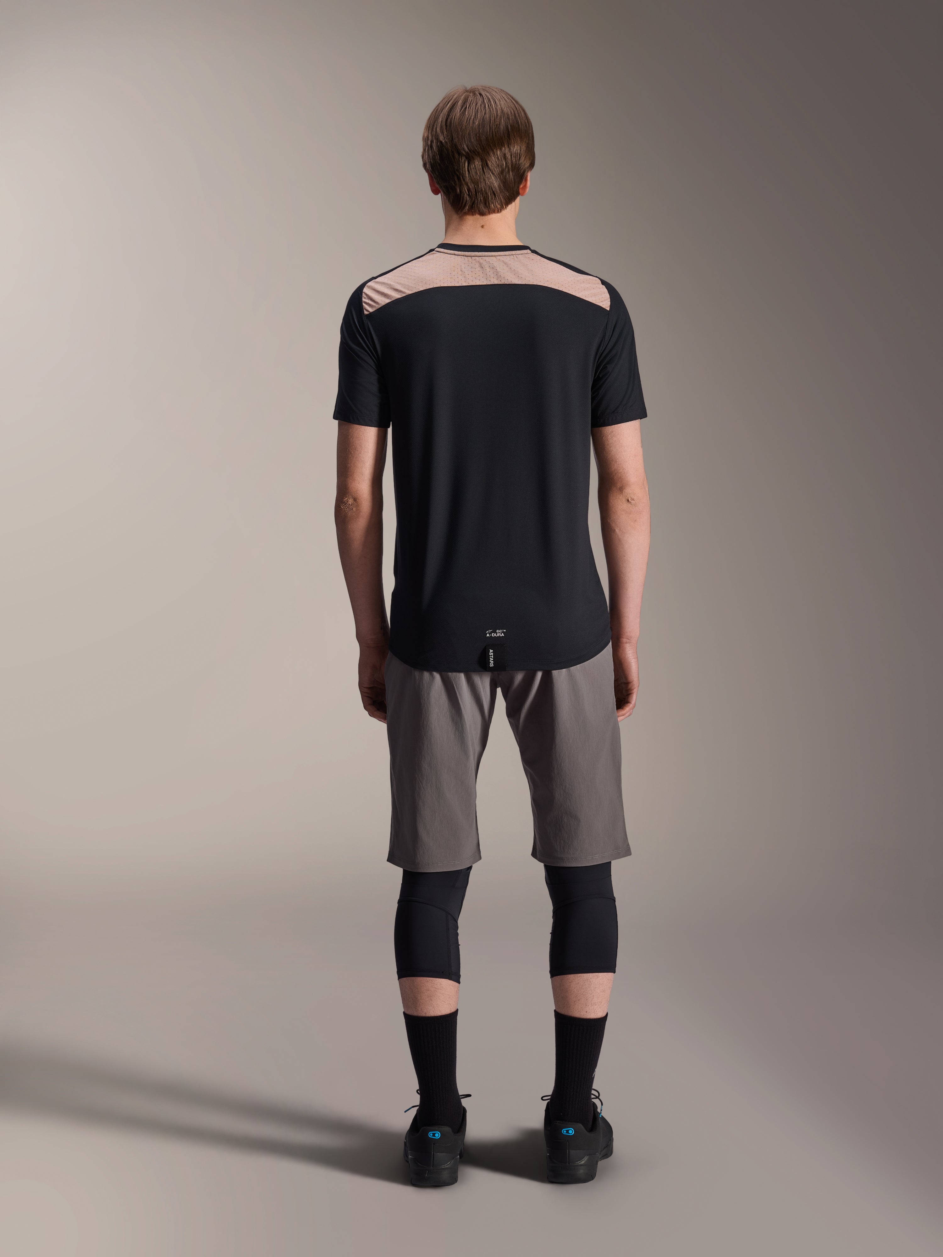 Alpinestars A-Dura Lipan Jersey - Manga corta, negro con panel superior de la espalda en color tostado, vista trasera que muestra la cola más larga, usado con pantalones cortos de MTB grises y rodilleras negras, ropa de alto rendimiento para bicicleta de montaña