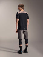 Camiseta de manga corta Alpinestars A-Dura Lipan en negro, vista trasera en modelo que muestra el panel de malla perforada marrón claro en la parte superior de la espalda, combinada con pantalones cortos de ciclismo de montaña grises y rodilleras negras para un conjunto completo de trail.
