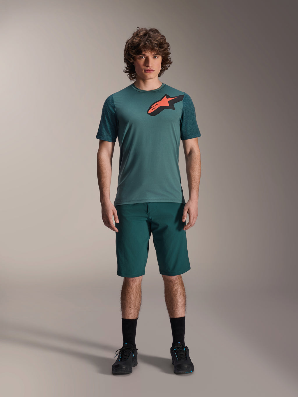 Alpinestars A-Dura Shorts en Dark Pine, pantalones cortos de ciclismo de montaña para hombre, mostrados en un modelo con una camiseta de manga corta verde azulada a juego con un logo de estrella rojo y negro, calcetines negros y zapatillas de ciclismo, ropa deportiva casual de MTB