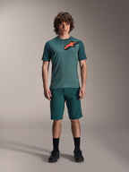 Alpinestars A-Dura Shorts en Dark Pine, pantalones cortos de ciclismo de montaña para hombre, mostrados en un modelo con una camiseta de manga corta verde azulada a juego con un logo de estrella rojo y negro, calcetines negros y zapatillas de ciclismo, ropa deportiva casual de MTB