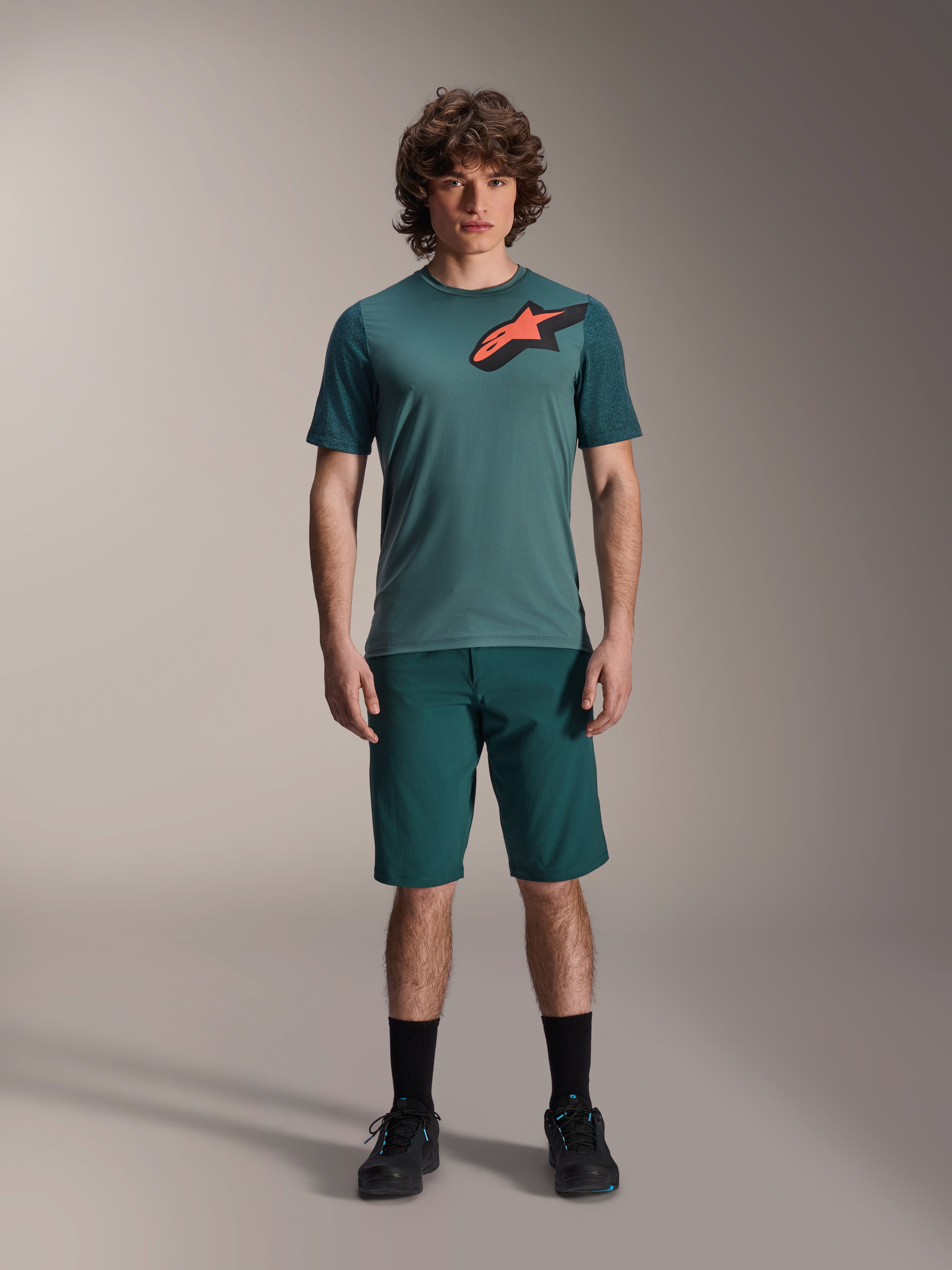 Alpinestars A-Dura Shorts en Dark Pine, pantalones cortos de ciclismo de montaña para hombre, mostrados en un modelo con una camiseta de manga corta verde azulada a juego con un logo de estrella rojo y negro, calcetines negros y zapatillas de ciclismo, ropa deportiva casual de MTB