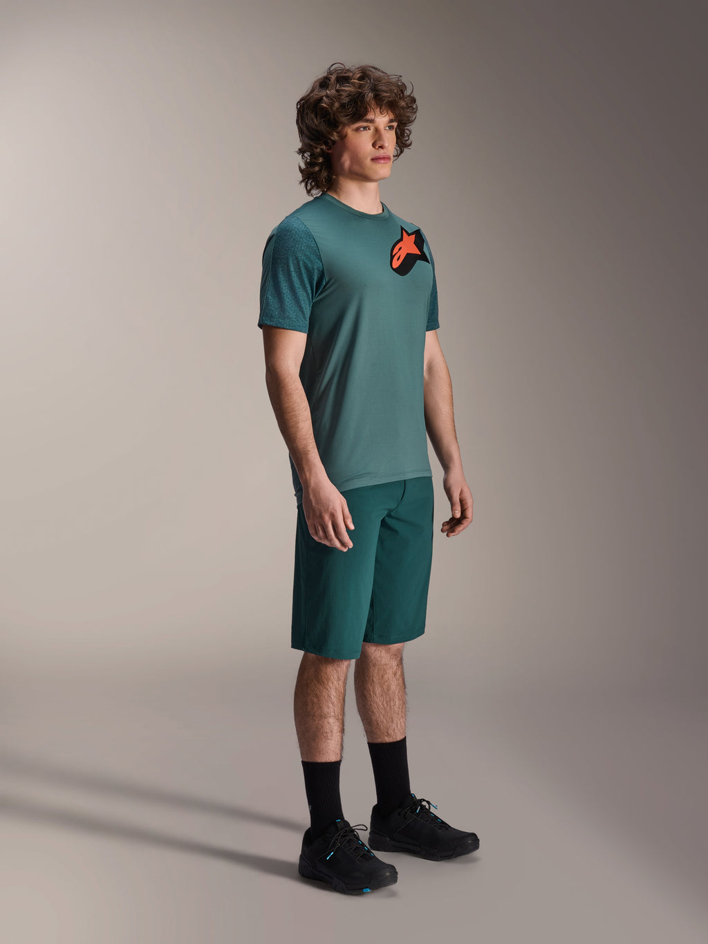 Alpinestars A-Dura Lipan Jersey, camiseta de MTB de manga corta, verde Silver Pine, con un logotipo Astars naranja y negro en el pecho, combinada con pantalones cortos verdes a juego y zapatillas de ciclismo negras, estilo trail casual