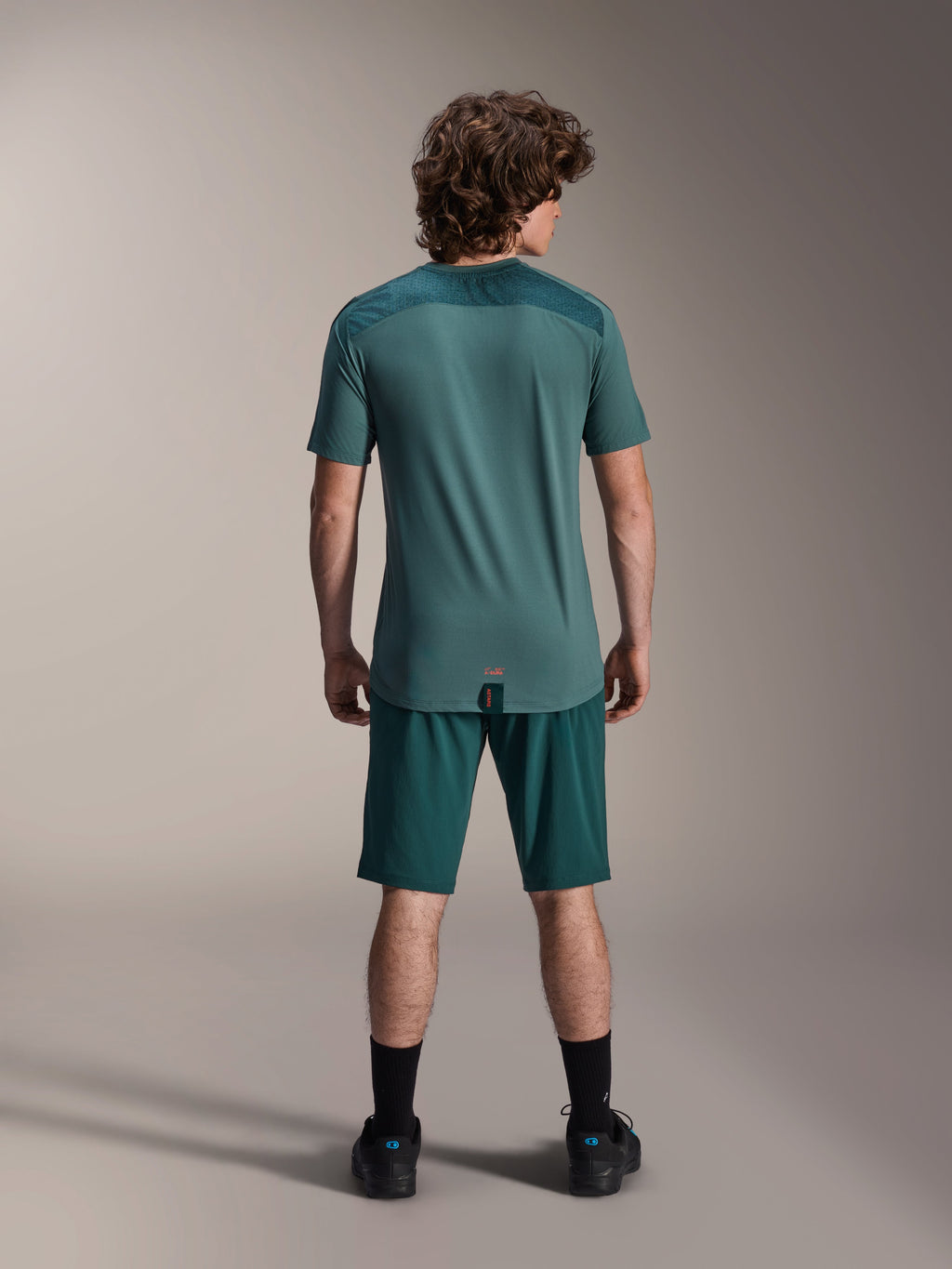 Jersey de manga corta Alpinestars A-Dura Lipan en Silver Pine, vista trasera que muestra los paneles de jacquard texturizado en los hombros y el dobladillo con cola más larga, usado con pantalones cortos de MTB verde oscuro a juego y calzado negro