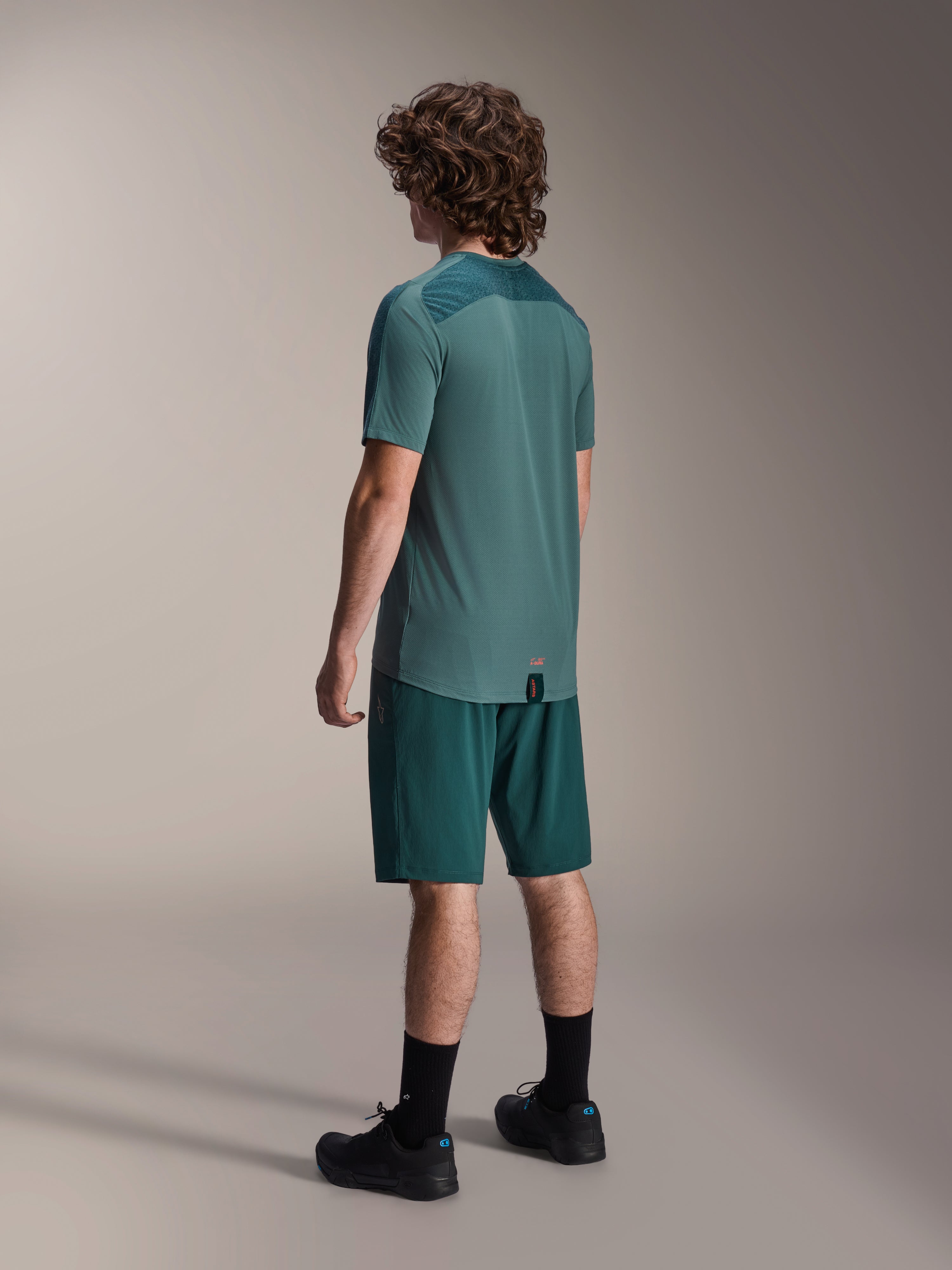 Modelo masculino vistiendo la camiseta de manga corta Alpinestars A-Dura Lipan en Silver Pine, visto desde atrás, combinada con pantalones cortos de ciclismo de montaña verde oscuro a juego, calcetines y zapatillas negras; cuenta con un panel superior de la espalda texturizado y un dobladillo trasero más largo para mayor cobertura.