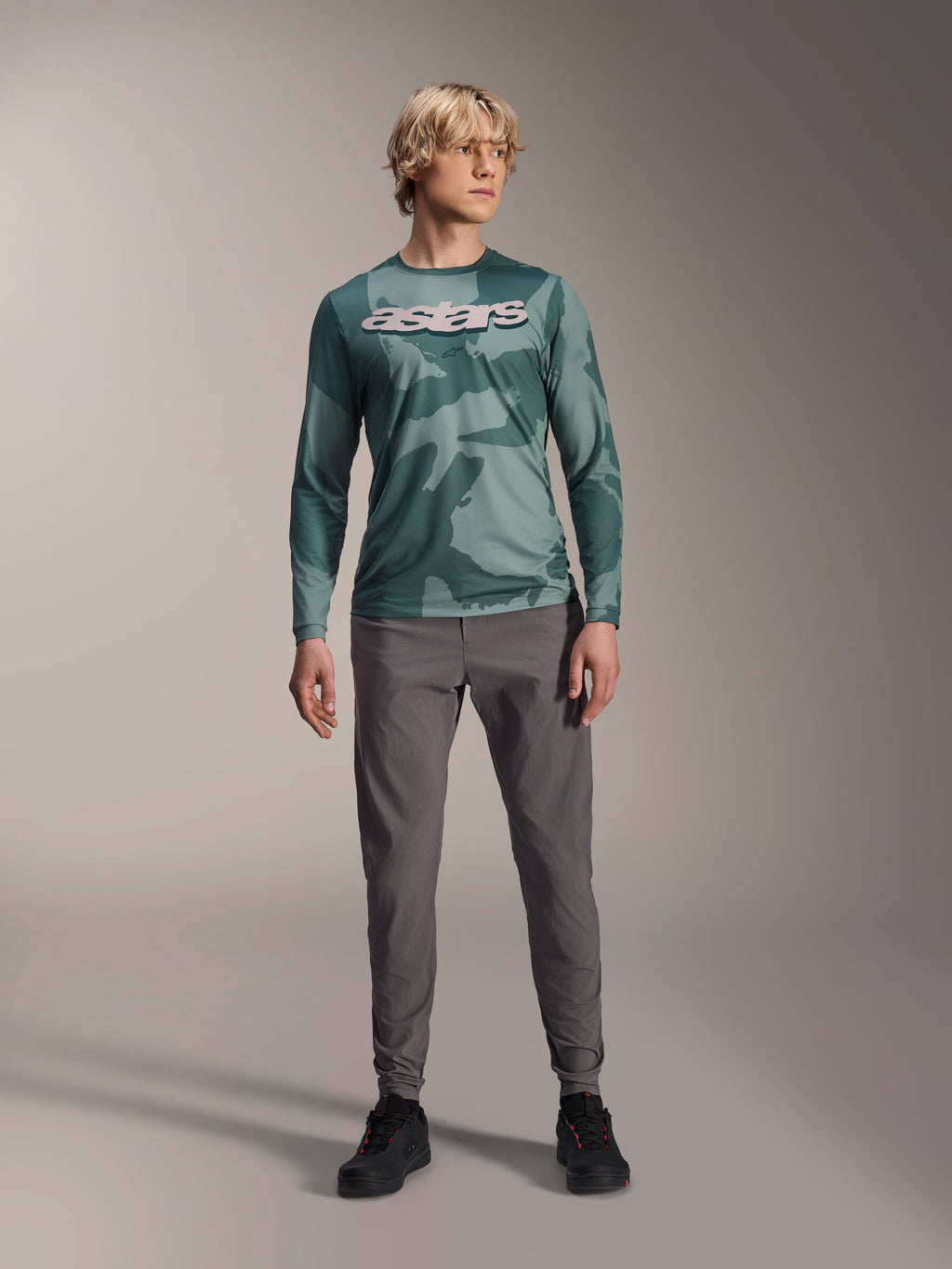 Jersey de manga larga Alpinestars A-Dura Hollow Camo, color Silver Pine Camo con patrón de camuflaje verde, logotipo "astars" grande en blanco en el pecho, combinado con pantalones técnicos grises y zapatillas de ciclismo negras, equipamiento de MTB de alto rendimiento