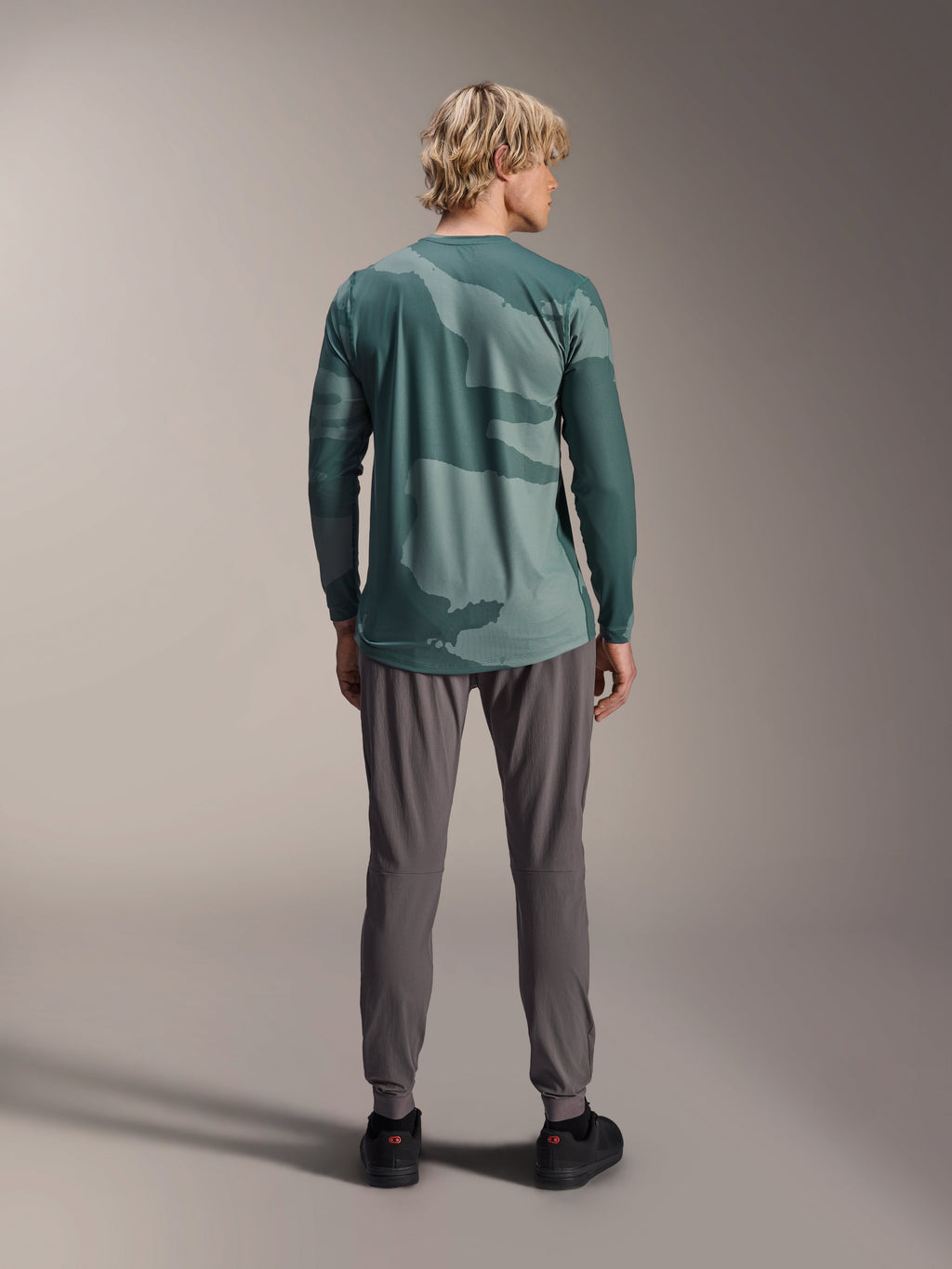 Alpinestars A-Dura Hollow Camo Jersey - Manga Larga, patrón de camuflaje verde Silver Pine Camo, modelo visto desde atrás con pantalones técnicos grises, ropa de alto rendimiento para bicicleta de montaña
