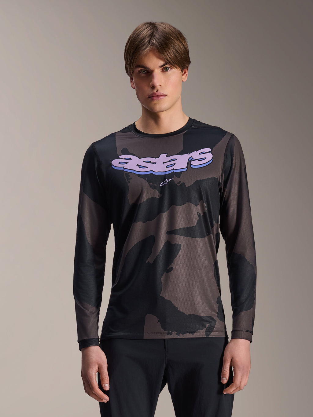 Alpinestars A-Dura Hollow Camo Long Sleeve Jersey, camiseta de manga larga para ciclismo de montaña, estampado de camuflaje negro y gris, gráfico 'astars' morado en el pecho, usada por un modelo con pantalones negros, diseño casual de alto rendimiento