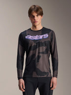 Alpinestars A-Dura Hollow Camo Long Sleeve Jersey, camiseta de manga larga para ciclismo de montaña, estampado de camuflaje negro y gris, gráfico 'astars' morado en el pecho, usada por un modelo con pantalones negros, diseño casual de alto rendimiento