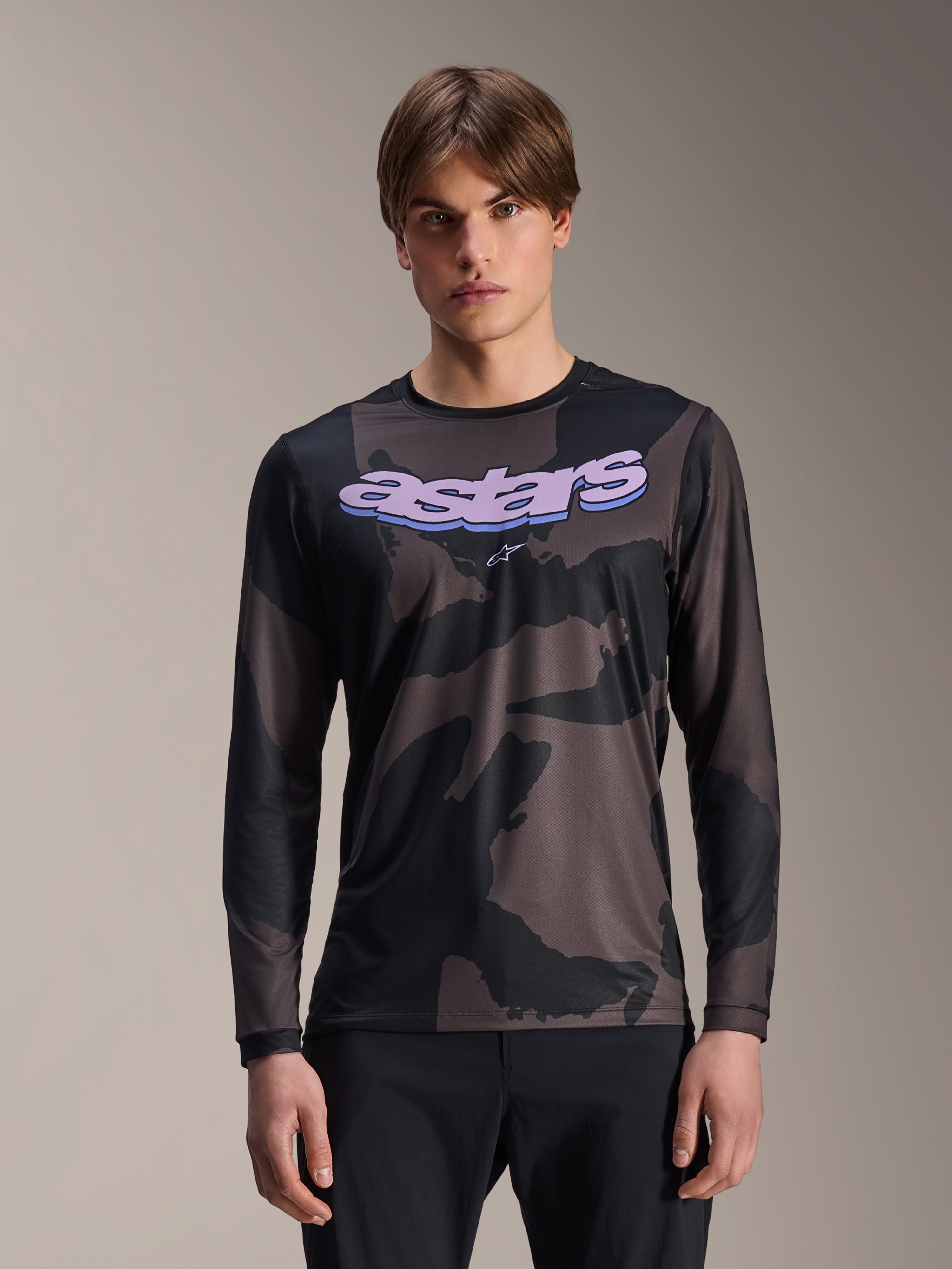 Alpinestars A-Dura Hollow Camo Long Sleeve Jersey, camiseta de manga larga para ciclismo de montaña, estampado de camuflaje negro y gris, gráfico 'astars' morado en el pecho, usada por un modelo con pantalones negros, diseño casual de alto rendimiento