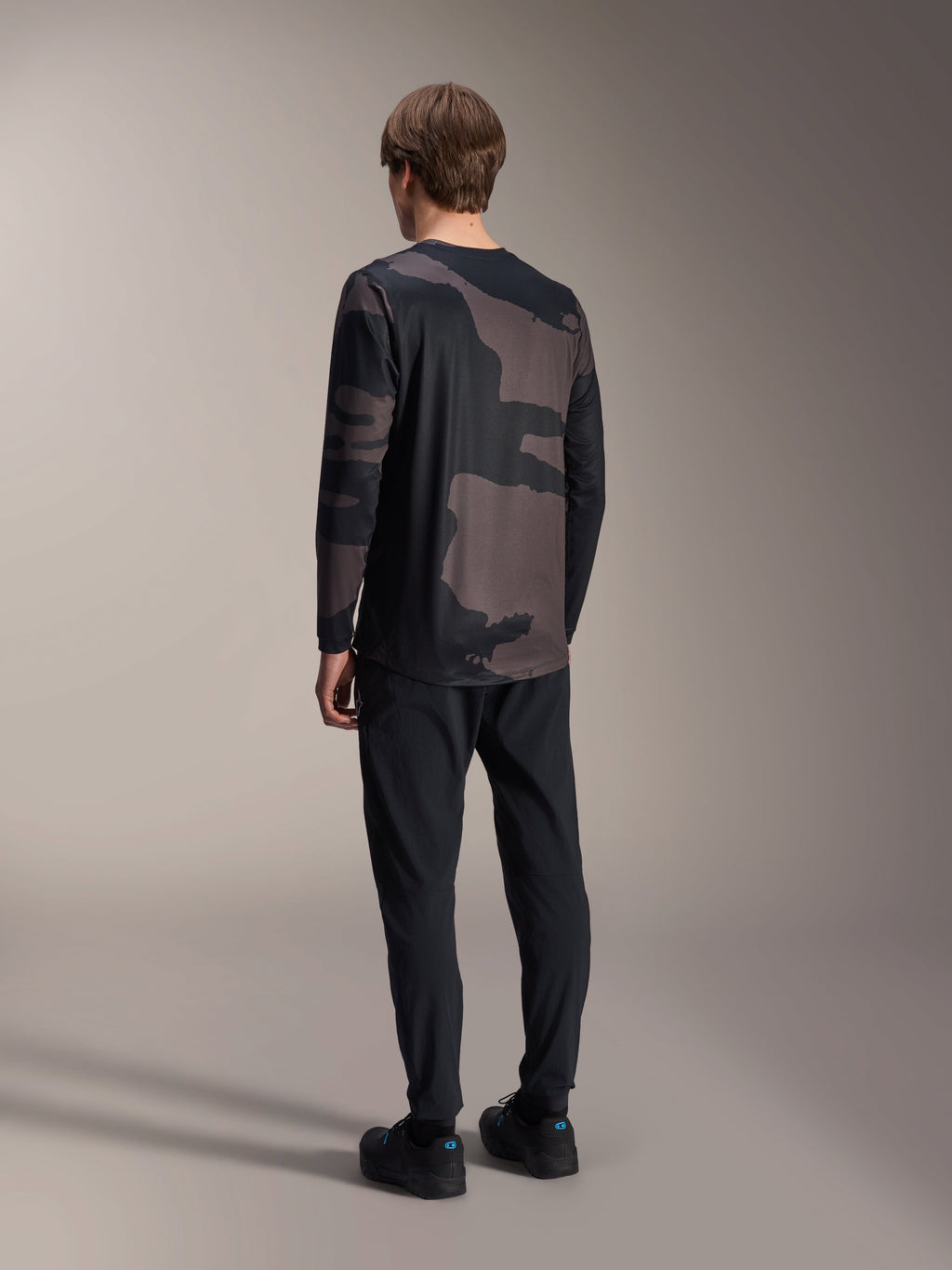 Alpinestars A-Dura Hollow Camo Jersey de manga larga, camuflaje negro, vista trasera del modelo, camiseta técnica de ciclismo de montaña con dobladillo trasero más largo, combinada con pantalones negros y zapatillas de ciclismo, equipamiento casual para todoterreno