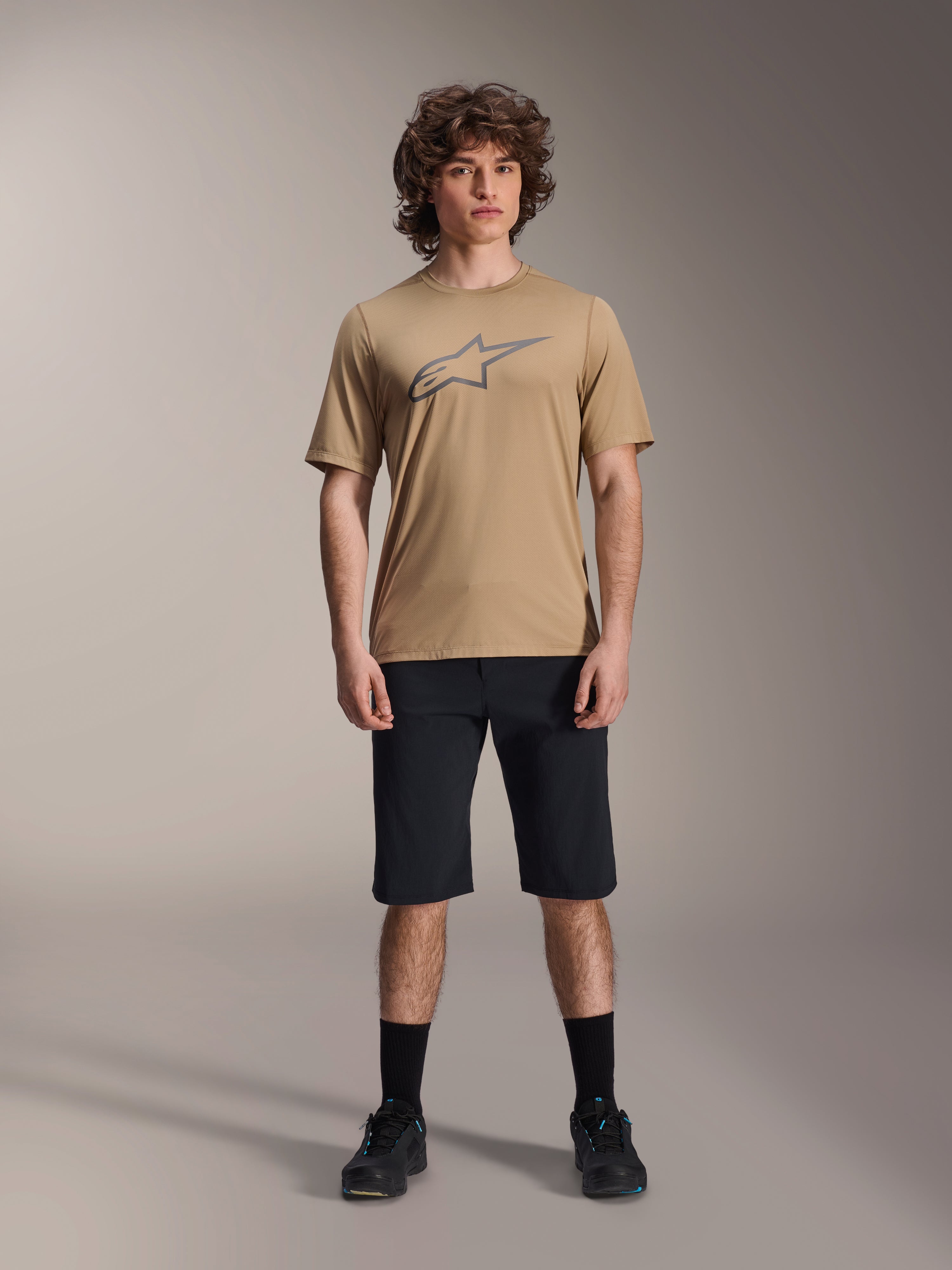 Model wearing Alpinestars A-Dura SS Jersey in tan and A-Dura Shorts in black, featuring a grey star logo on the chest, paired with black socks and mountain biking shoes, complete MTB riding apparel in a studio setting.
