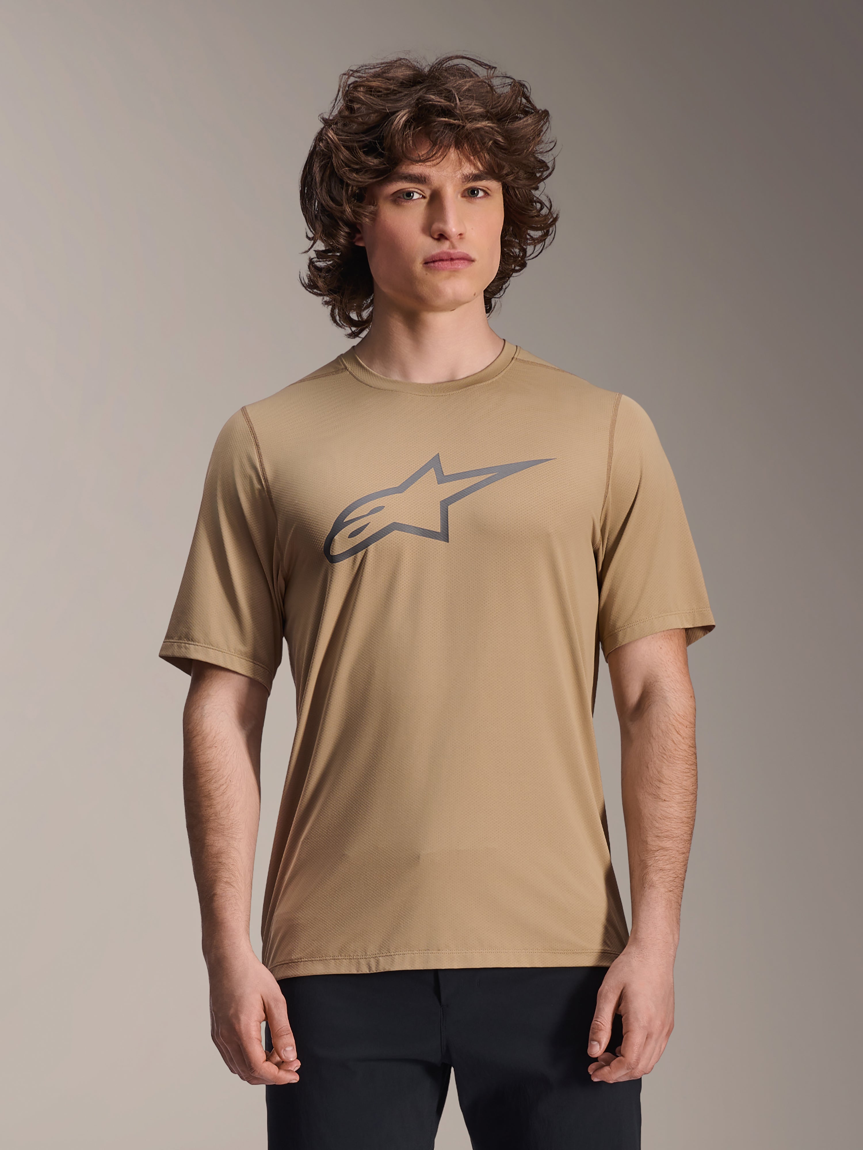 Alpinestars A-Dura Astar Short Sleeve Jersey, marrón claro, usado por un modelo masculino con pantalones negros, logotipo de estrella Alpinestars en gris en el pecho, jersey casual de ciclismo de montaña para trail