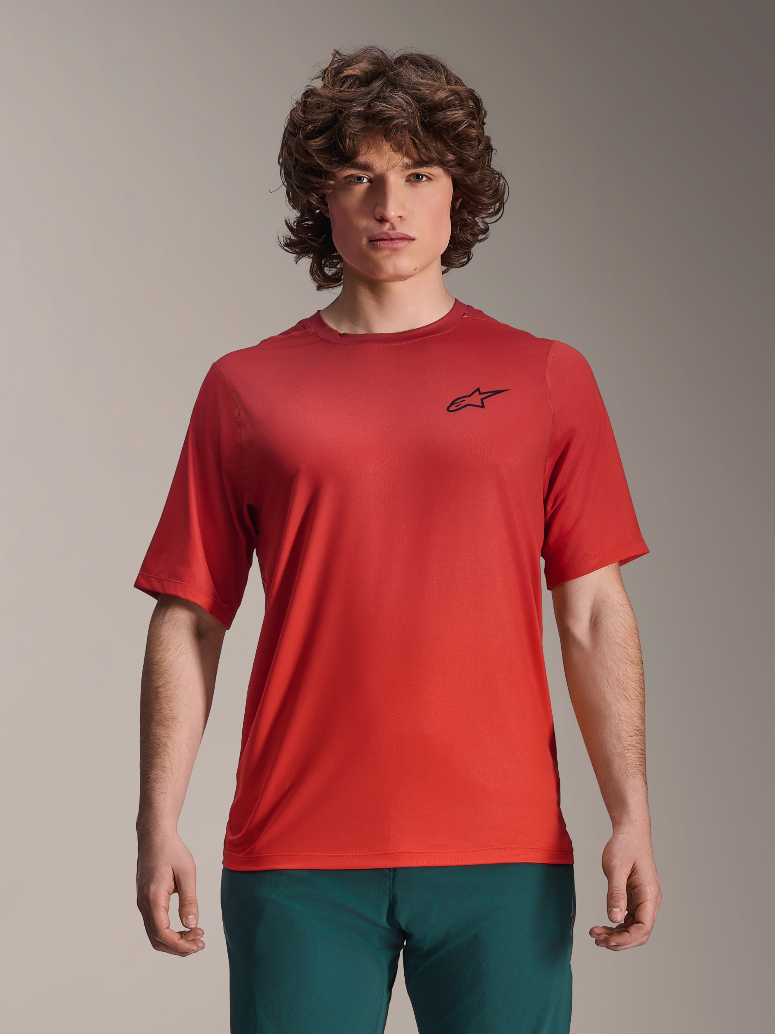 Alpinestars 2025 A-Dura Rise Jersey, camiseta de MTB de manga corta, color Spicy Orange, usada por un hombre, con un logotipo negro de Astars en el pecho, combinada con pantalones verde oscuro, ropa técnica casual para ciclismo de montaña